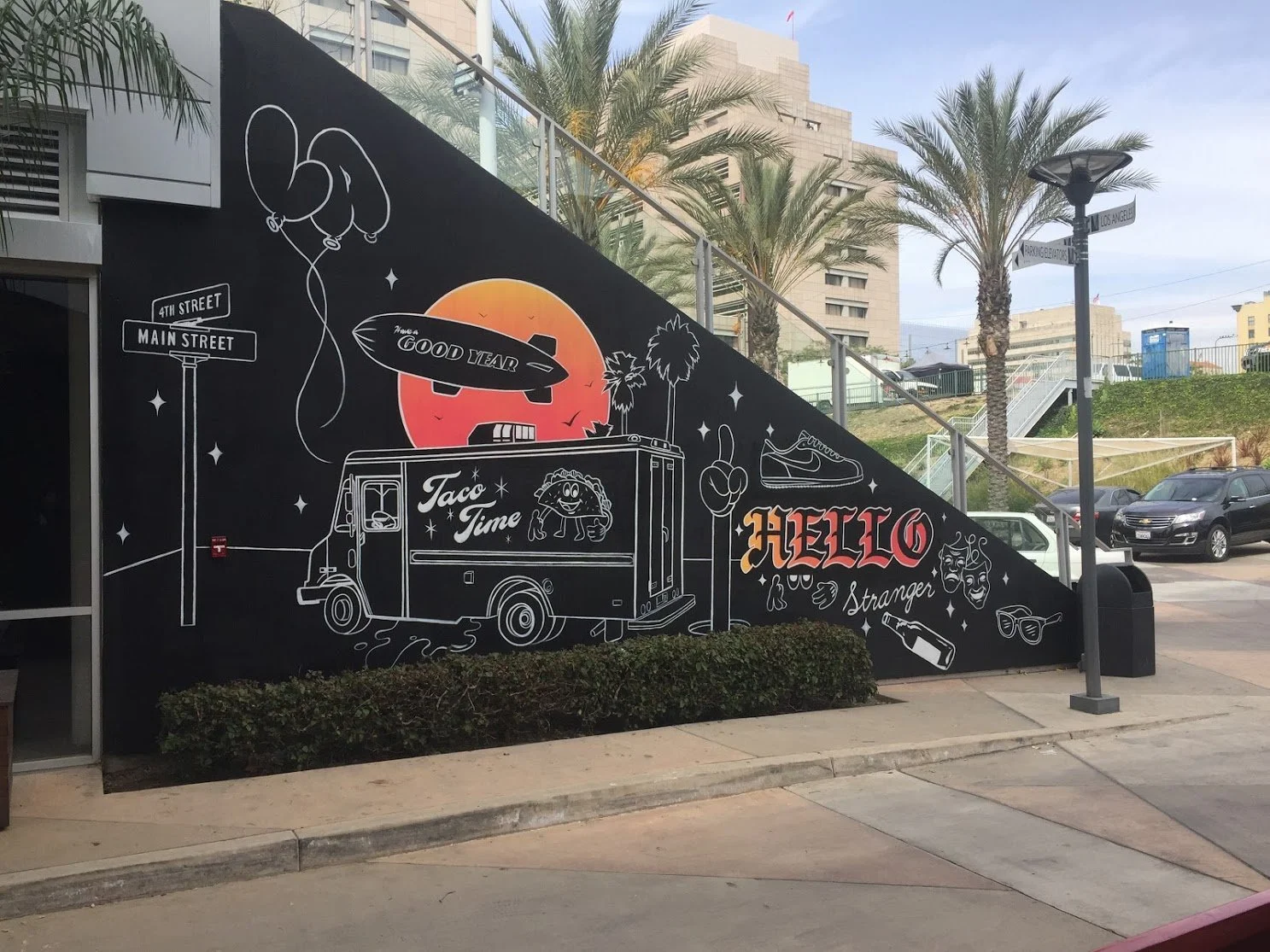 A black mural on an outdoor wall featuring white illustrations of a taco truck, food items, palm trees, a sunset, and various decorative elements with the words 'Hello Stranger' and 'Taco Time'.