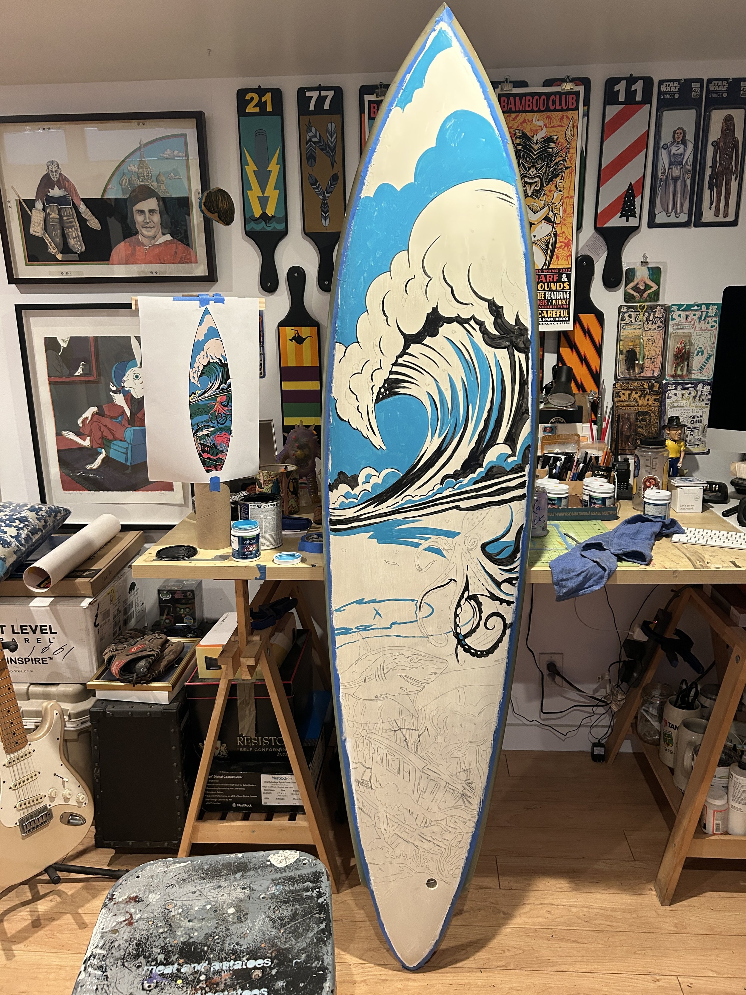 This image shows a workspace with a surfboard in progress, featuring a large wave design and pencil sketches. The wall in the background has various vintage-looking paddleboard and surf-themed signs, posters, and artwork. There are art supplies, a small lamp, a clothing item, and a computer on the desk. A guitar and several boxes are also visible.