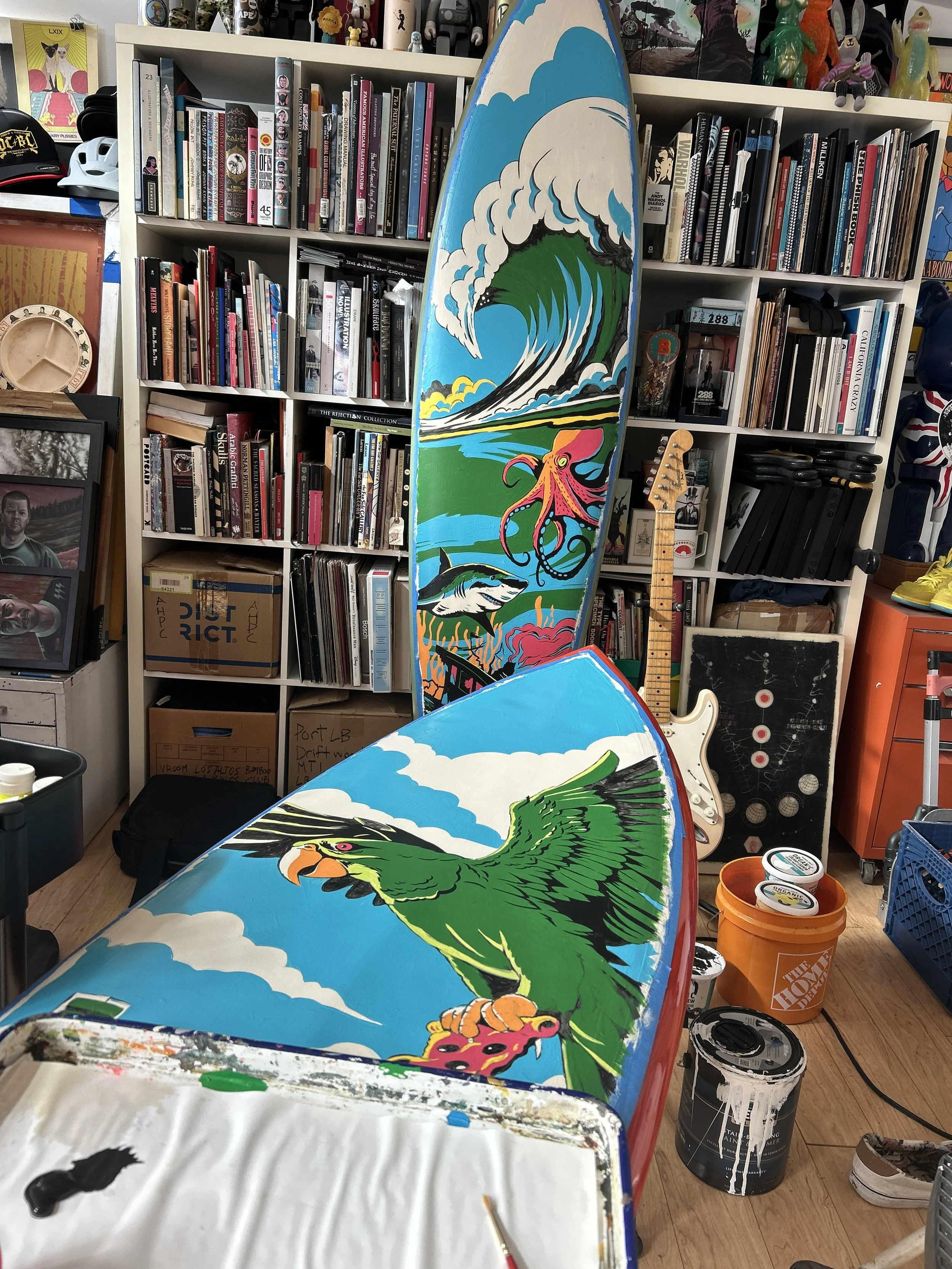 A colorful surfboard featuring a vibrant illustration of a parrot and a wave is in the foreground of a cluttered room filled with books, paintings, musical instruments, and various art supplies.