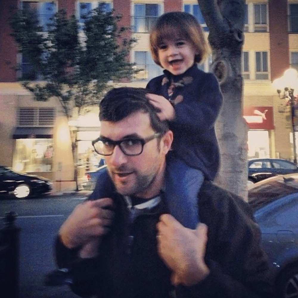 A man with glasses carrying a young girl on his shoulders in an outdoor urban setting at dusk, with trees and buildings in the background.