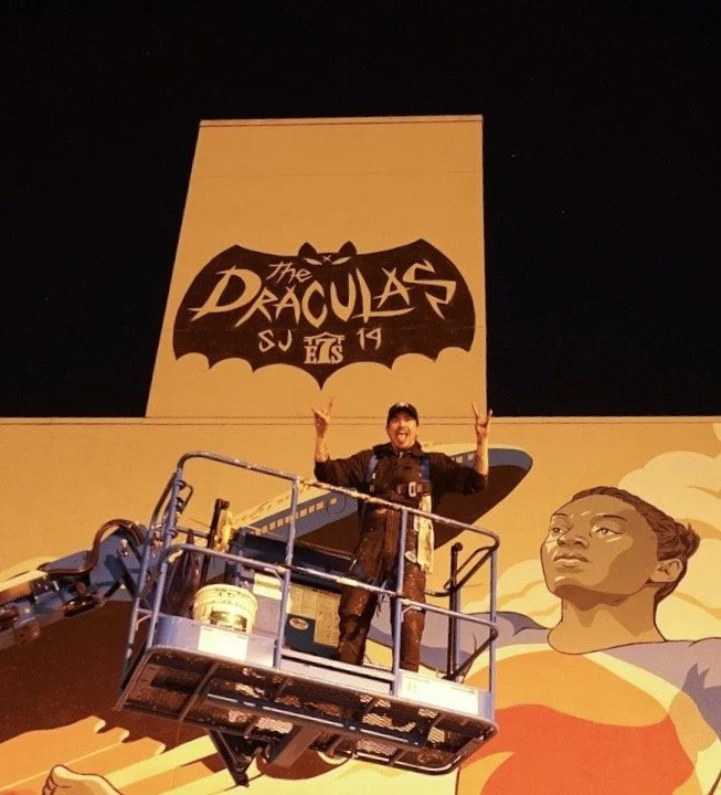 Person standing on a lifted platform in front of a mural advertising 'The Draculas' with a bat silhouette and a woman painted on the wall.