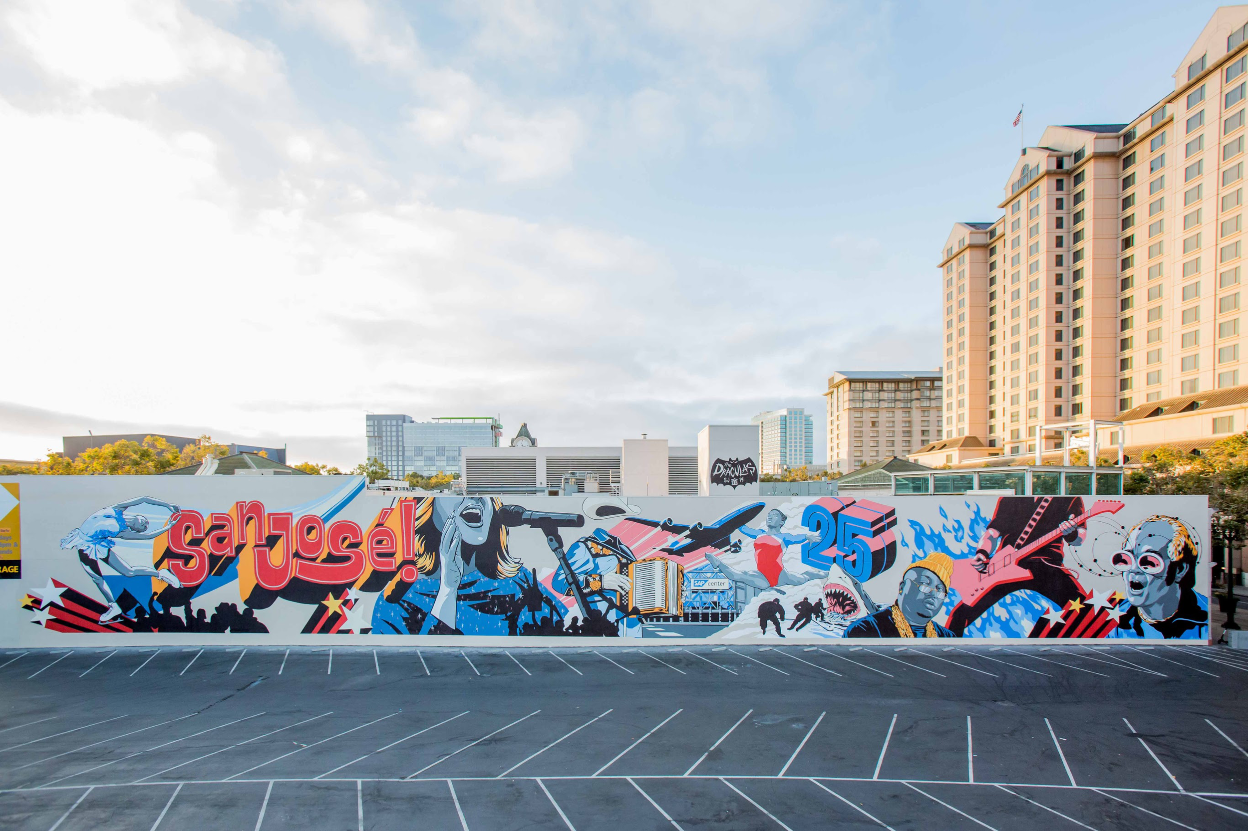 Colorful mural on a wall with various characters and text, including 'sanjose!', in front of tall buildings and a blue sky.