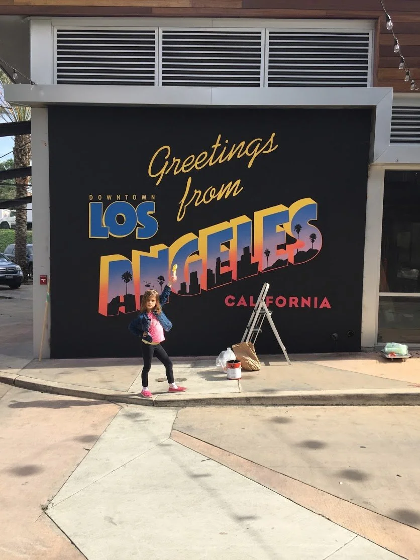 A young girl in pink shoes and a denim jacket standing in front of a large sign that says "Greetings from Los Angeles California." The sign features colorful lettering with a sunset gradient, palm trees, and cityscape silhouettes. There are some pain