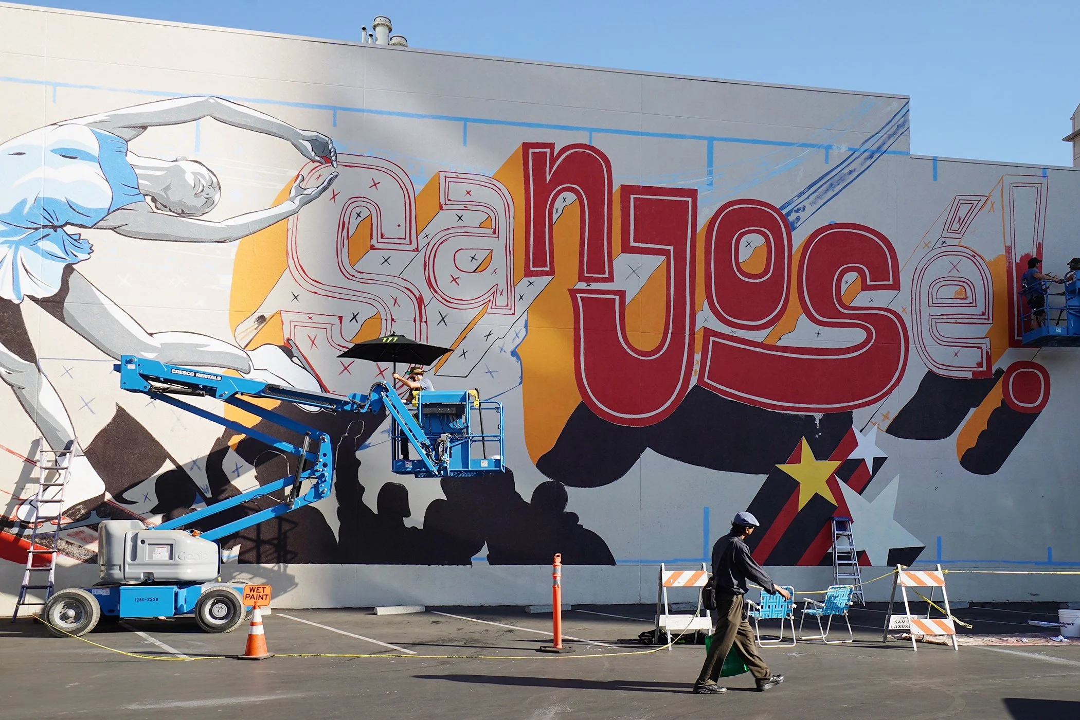 A large mural painting being created on the side of a building, featuring a stylized woman with blond hair in a white and blue outfit, reaching out with her hand. Several workers are using lifts and ladders to paint details, with cones and caution tape around the work area.