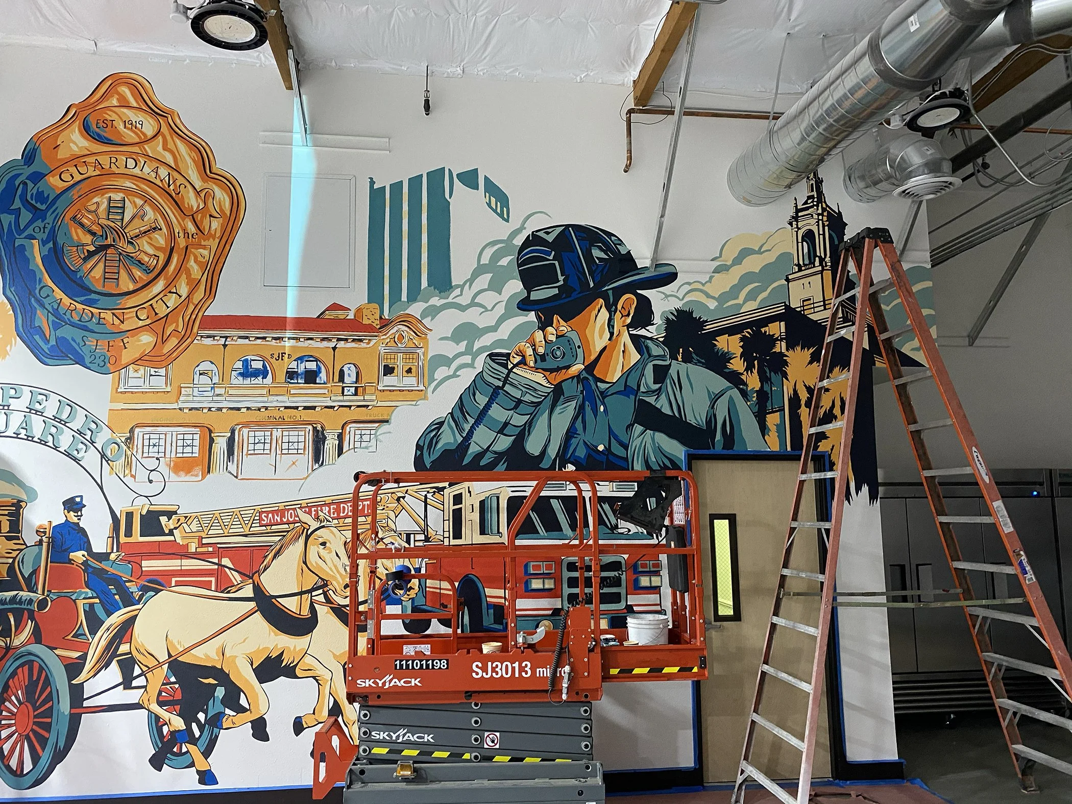 A colorful mural on a wall depicts a firehouse, fire trucks, and firefighters, with a firefighter holding a walkie-talkie. There is a ladder and a scissor lift in front of the mural, with some construction equipment and pipes visible on the ceiling.