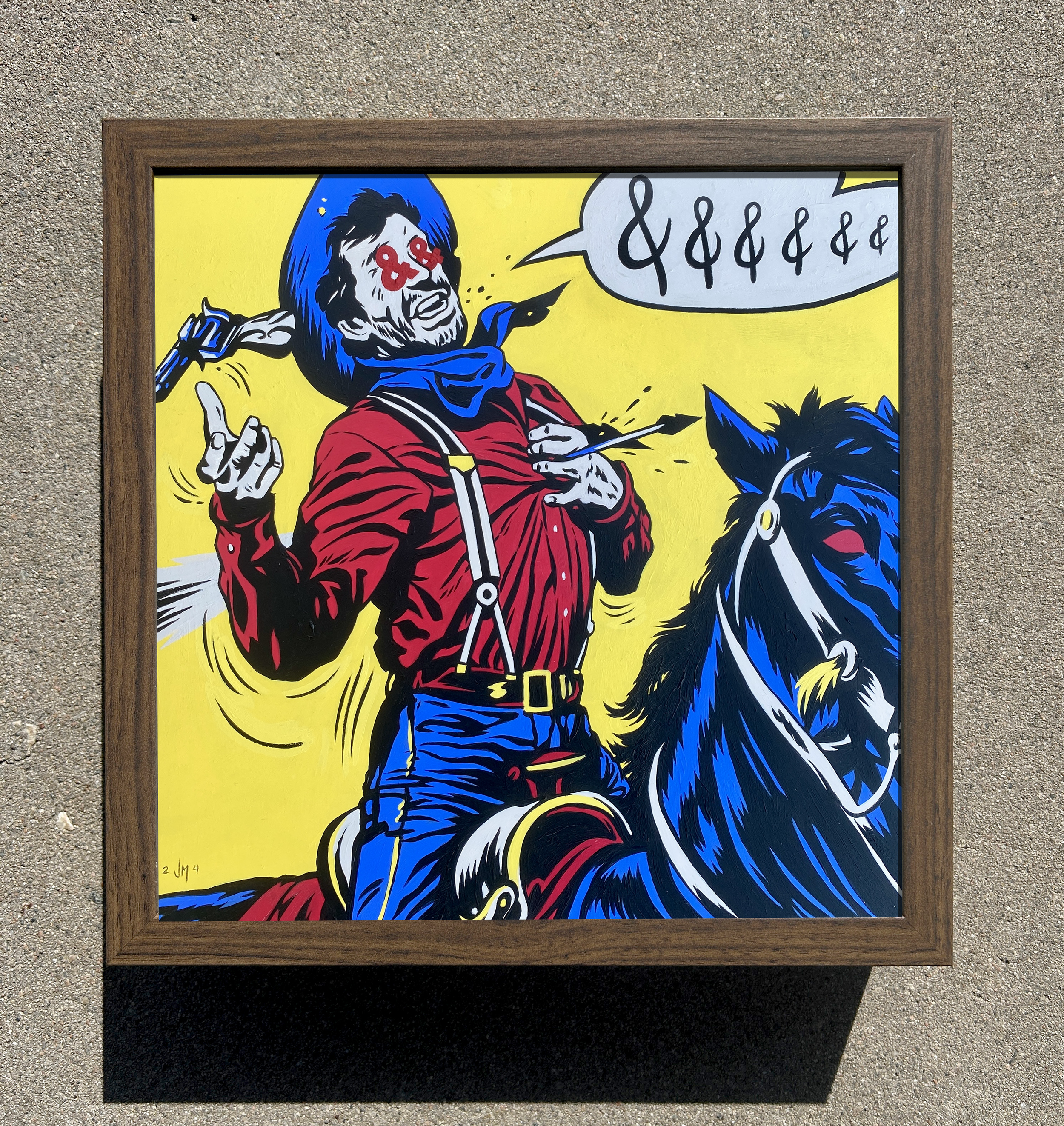 Pop art style painting of a man with dark hair, beard, and red shirt, pointing his finger upward, against a yellow background, with a speech bubble containing multiple ampersands. The painting is framed in wood and is placed on a rough outdoor surface.