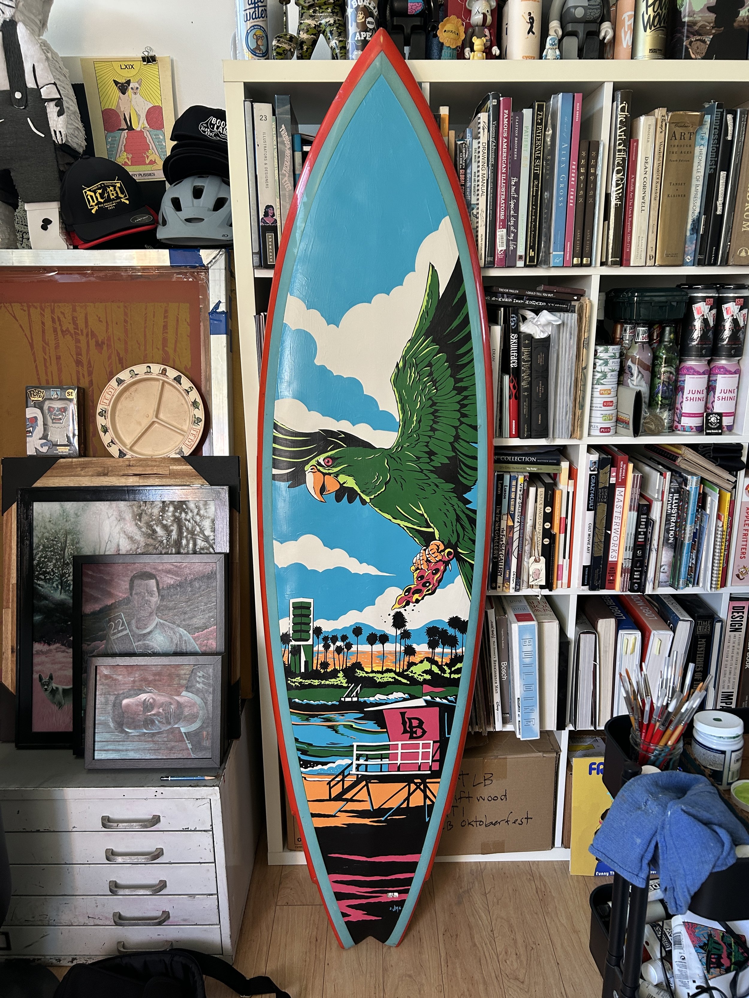 A colorful surfboard with a graphic of a green parrot holding a flamingo, standing in front of a landscape with palm trees and a sunset. It is placed in a room surrounded by bookshelves filled with books, hats, helmets, and various decorative objects.