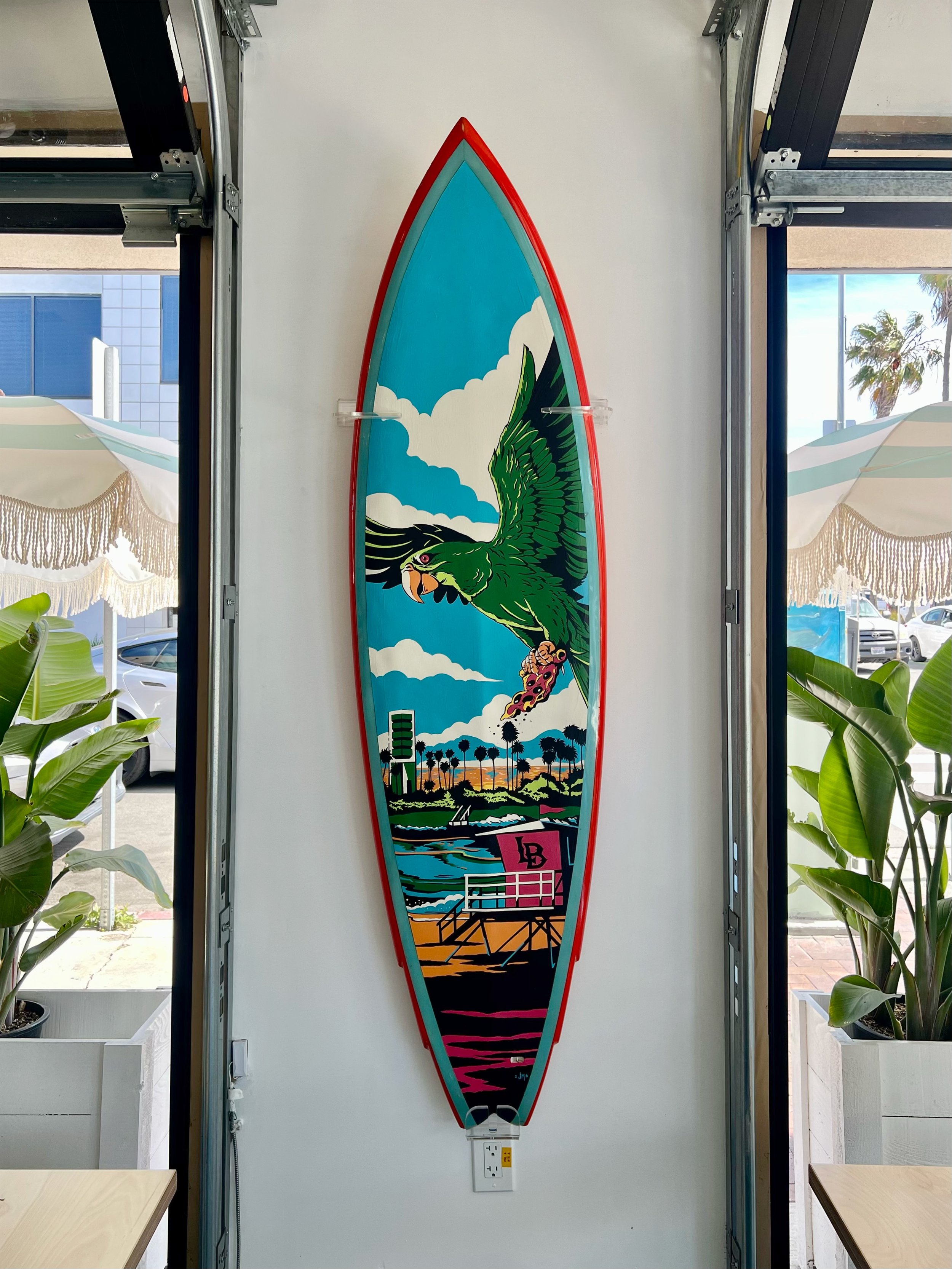 A colorful surfboard with a graphic of a green eagle flying over a landscape, mounted on a white wall between windows with plants nearby.