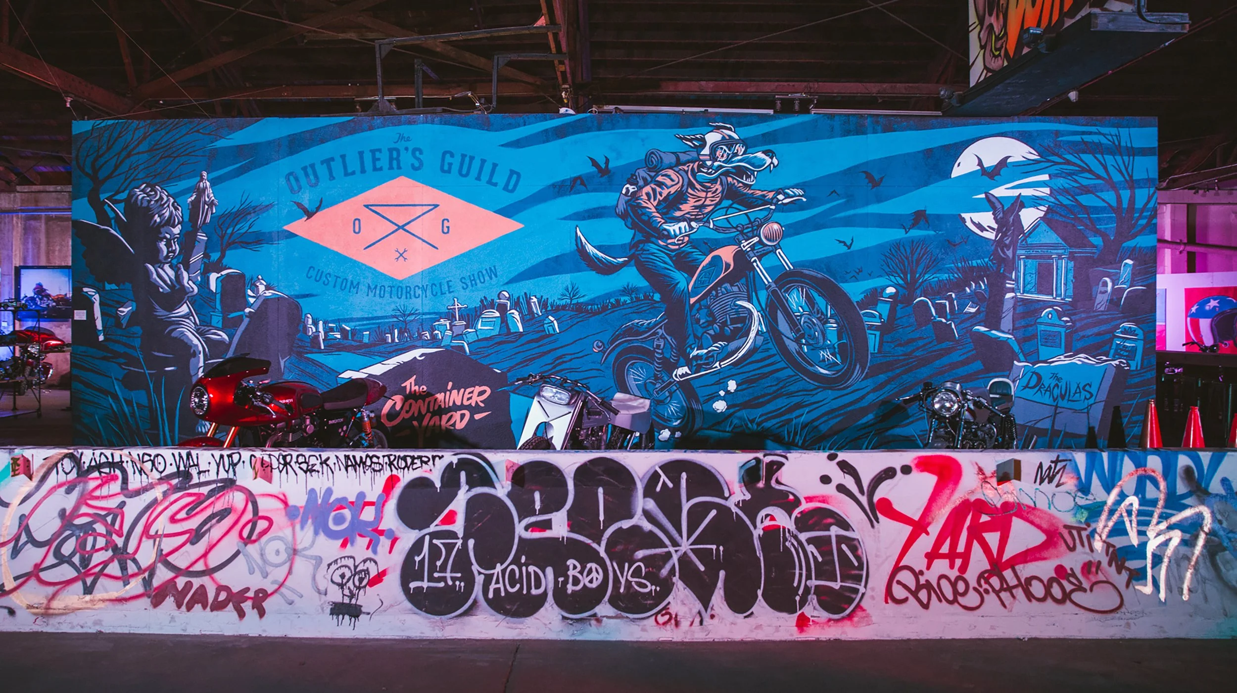 Colorful graffiti mural at an indoor motorcycle show, depicting a wolf riding a motorcycle against a blue background with trees, bats, and a moon. The mural has text that reads 'The Outlier's Guild' and 'Custom Motorcycle Show', with additional graff