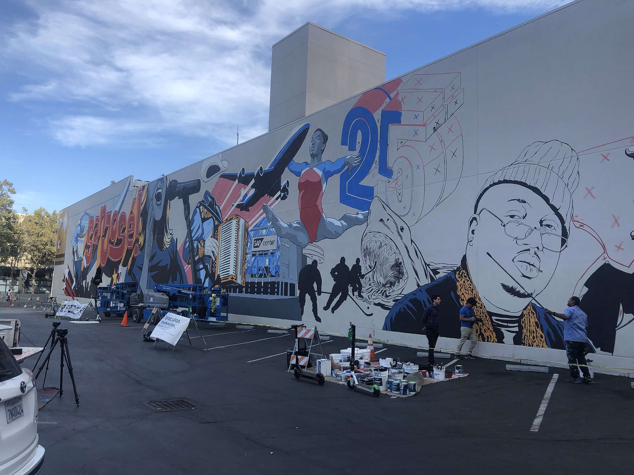 Large mural painting on a building wall celebrating 25 years, featuring diverse illustrations of people, a plane, and artistic elements with a lady holding a badminton racket, and various artists working on the mural.