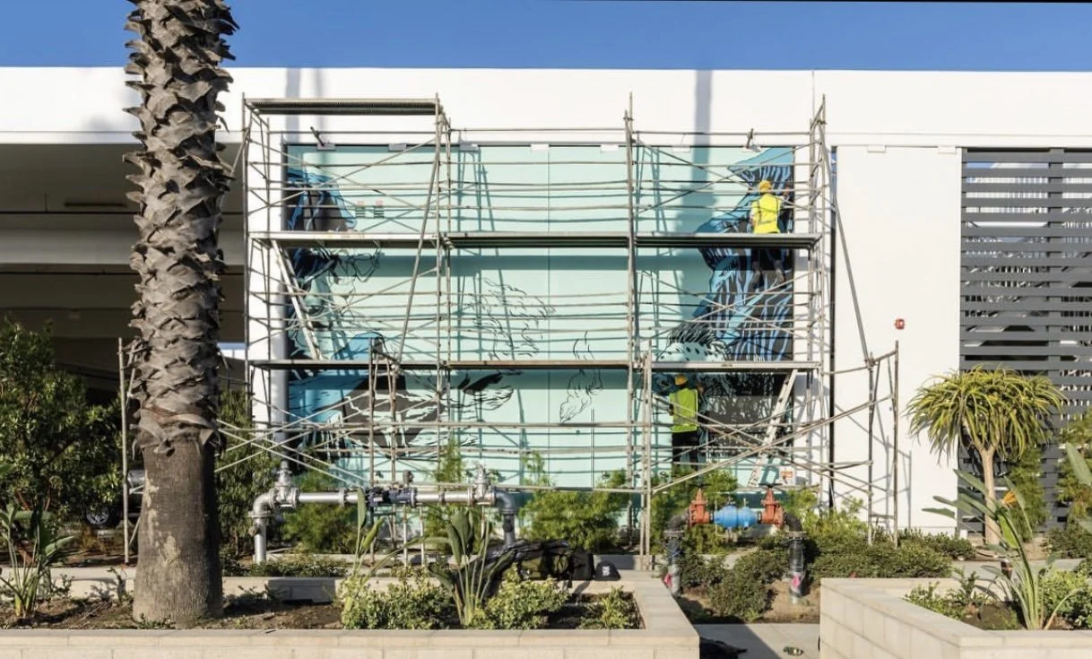 Workers installing or painting a mural on the exterior wall of a building, surrounded by scaffolding and plants.