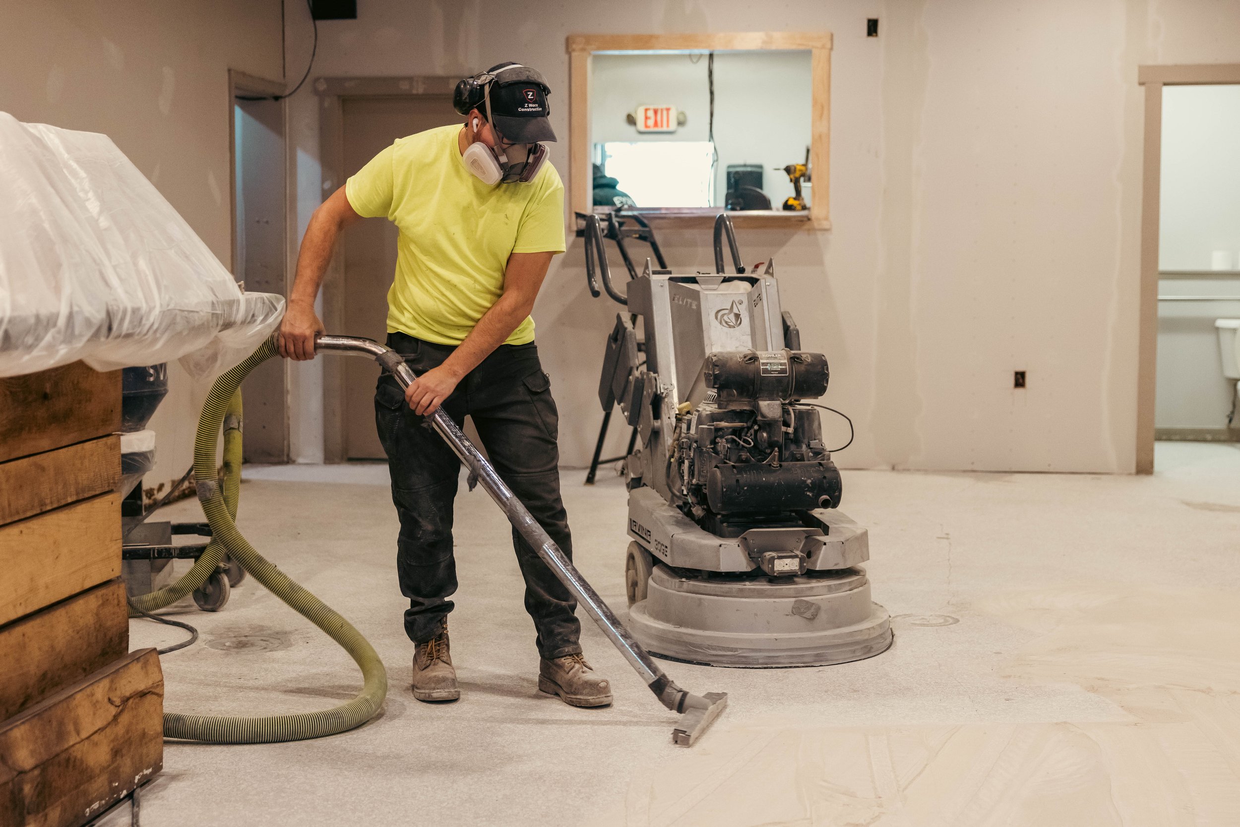 A construction worker wearing a yellow t-shirt, black pants, work boots, a cap, ear protection, and a dust mask is operating a concrete grinder in an unfinished room. The room has drywall walls, some tools, and an opening to another area with an exit sign above it.
