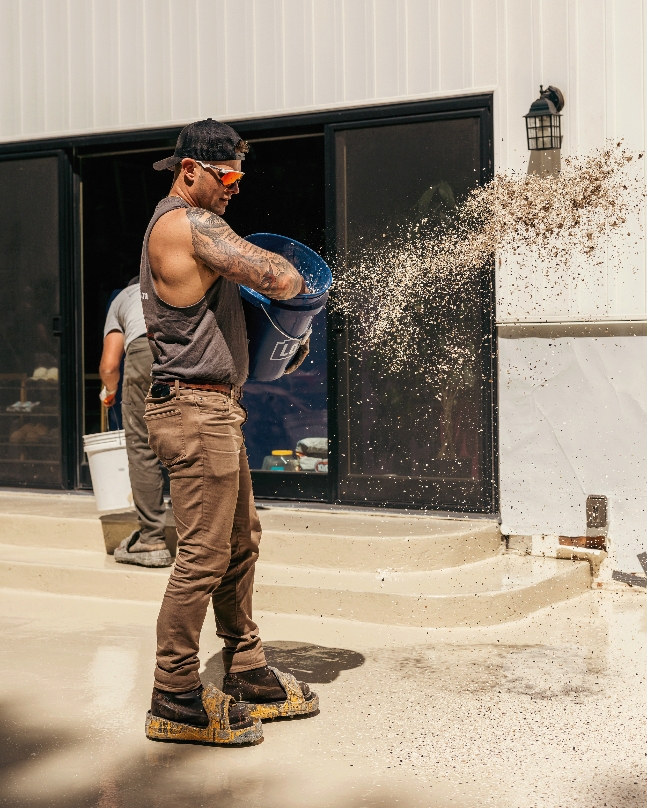 A man wearing sunglasses, a black cap backward, and a sleeveless shirt stands on a muddy ground, wearing platform shoes and large muddy sandals, as he pours a bucket of wet cement or concrete. There is another person partially visible in the background near a building with white and black exterior walls and a lantern-style light fixture.