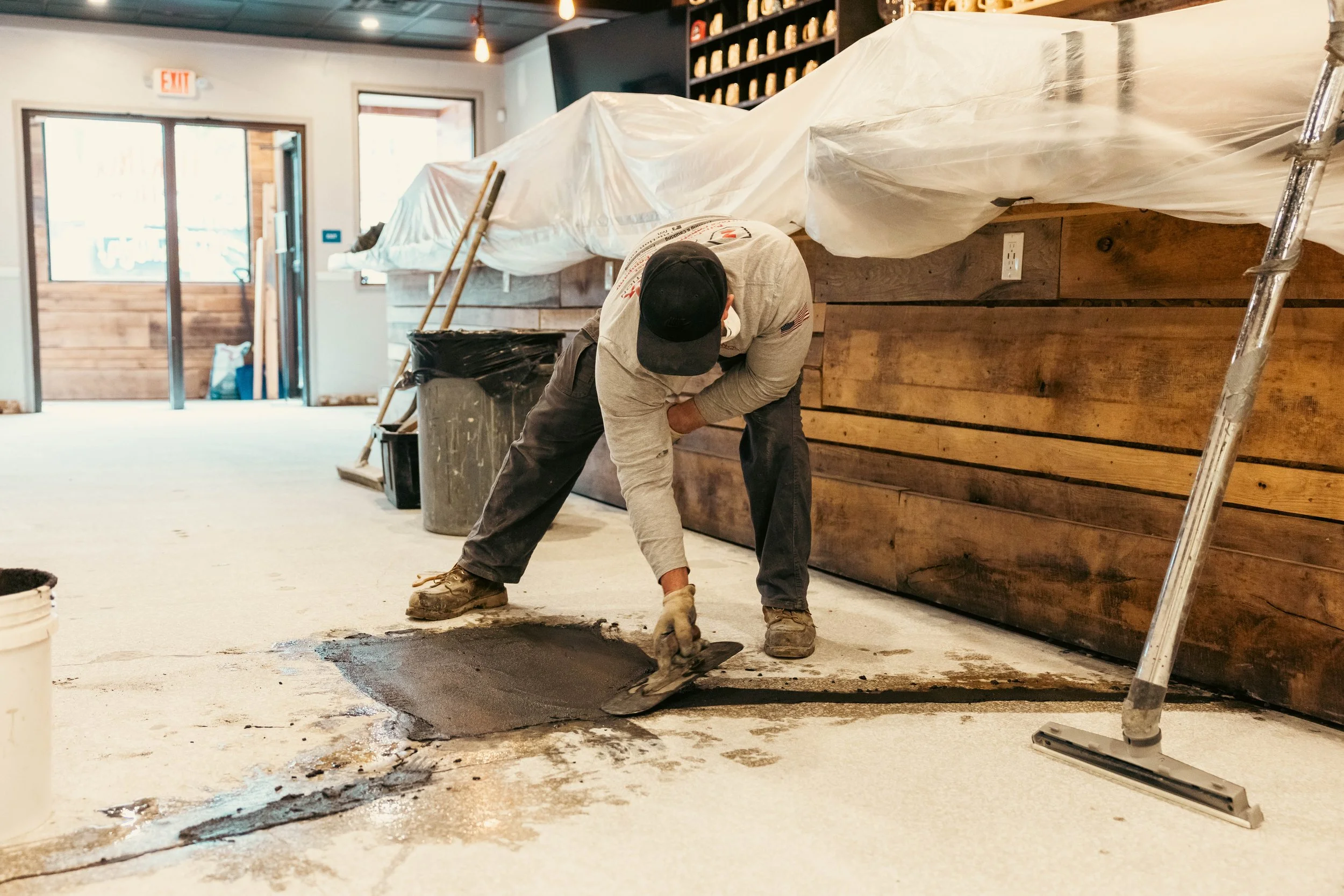 A person with a black cap and gloves is working on fixing a hole or crack in a wooden floor. The person is bent over and appears to be spreading or smoothing something dark on the floor. The space looks like a restaurant or cafe under renovation, with exposed light bulbs, wooden wall decor, and a doorway with glass panels in the background.