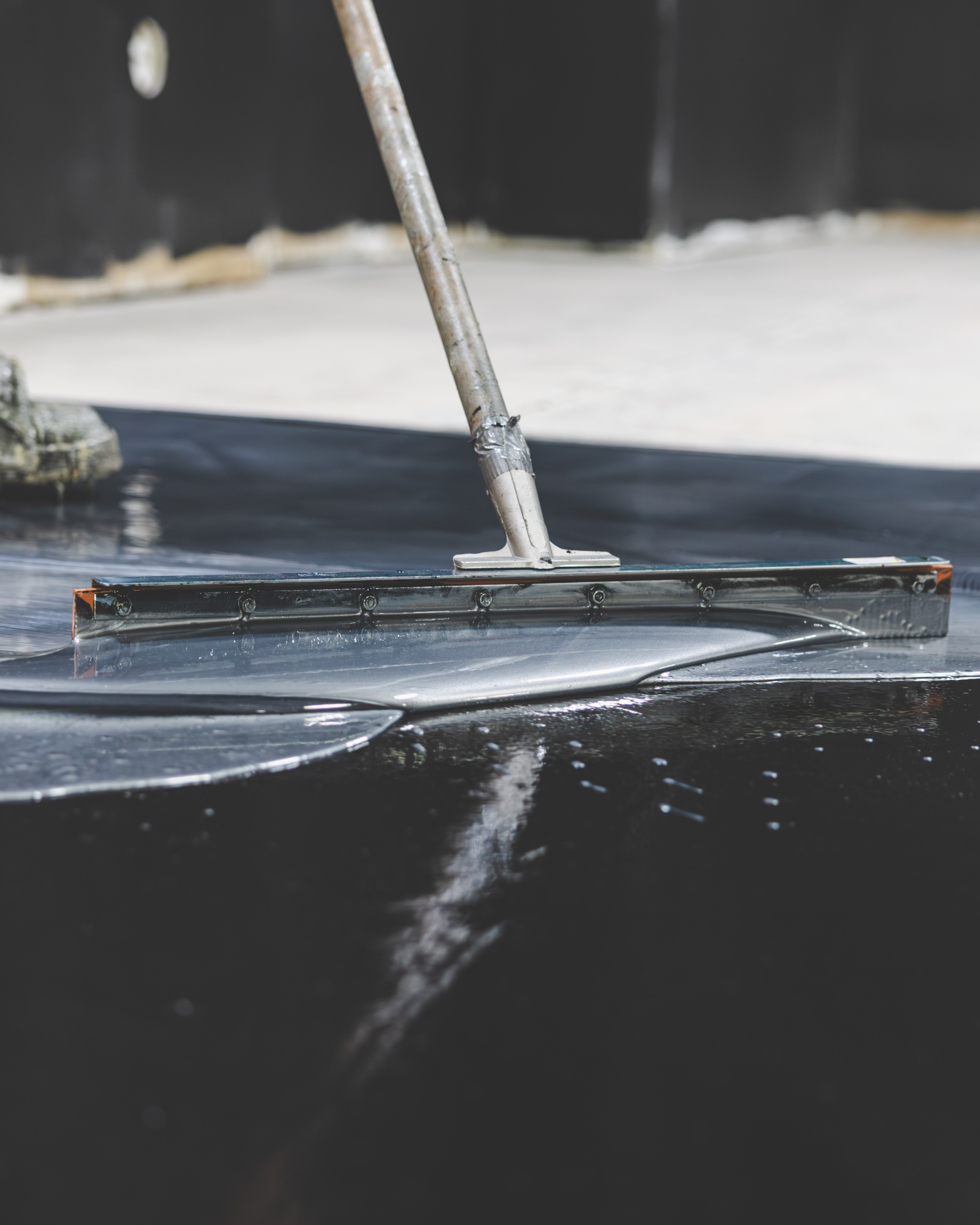 Close-up of a metal tool, likely a squeegee, being used to smooth or apply material on a dark surface, possibly a vehicle or a window.