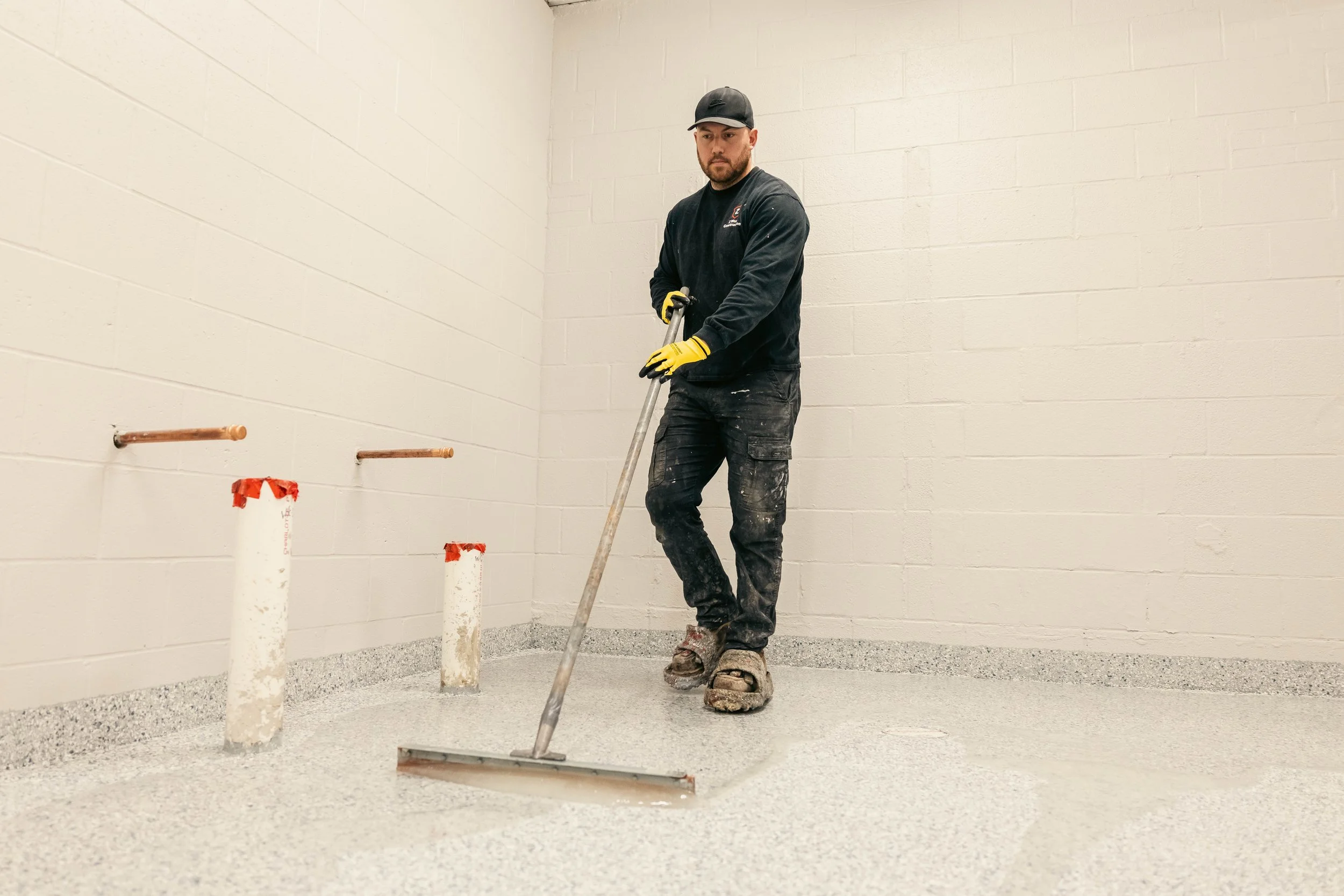 A man wearing work gloves, a black baseball cap, and black work clothes is using a tool to smooth fresh concrete on a floor. The room has plain white cinderblock walls and has pipes protruding from the wall, covered with red tape.