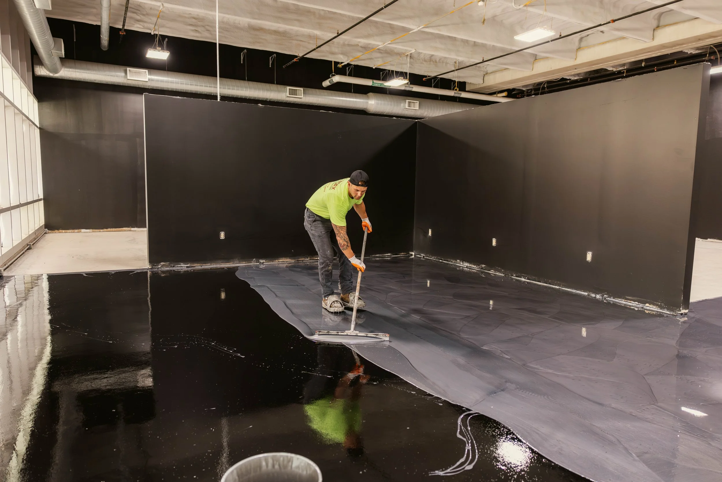 A worker in a lime-green shirt and black cap spreads black paint or coating on a floor with a roller in an indoor space with exposed ceiling pipes and a black wall.