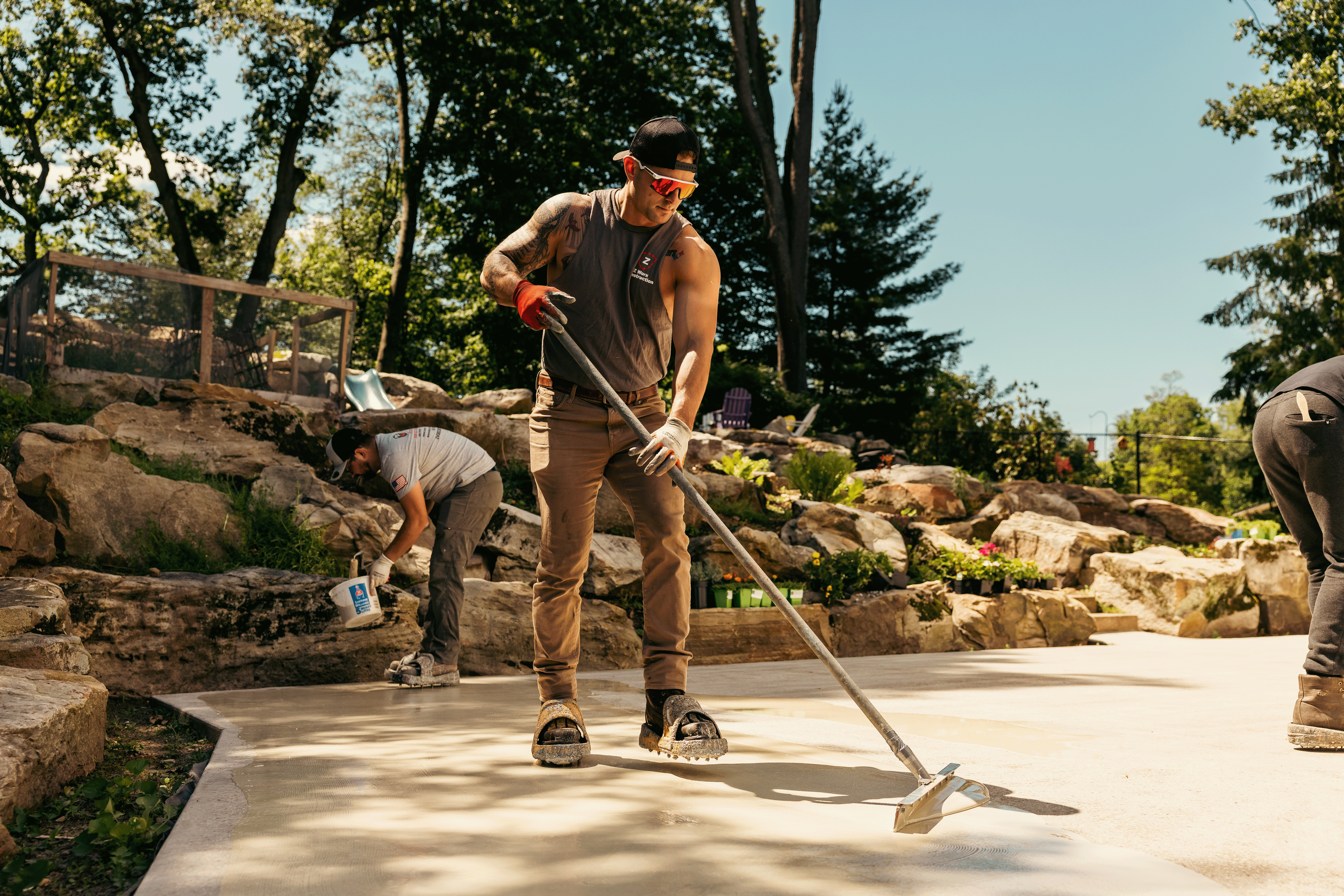A man wearing sunglasses, a backwards cap, a sleeveless shirt, and gloves is smoothing wet concrete with a drag squeegee. Two other people are working on the concrete in the background, with one using a bucket. The scene is outdoors, surrounded by trees and rocks in a landscaped yard.