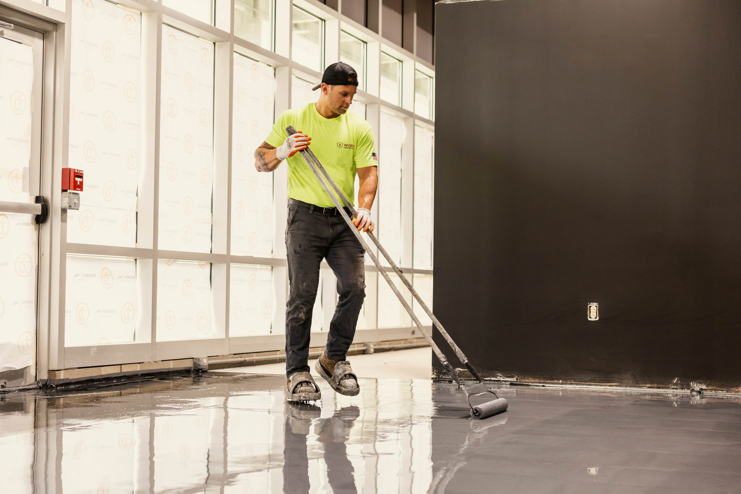 A man in a neon yellow T-shirt, black pants, and sandals is using a roller to spread a coating on a floor in a construction area. The construction site has unfinished walls with window panes and a red fire alarm box on the wall.