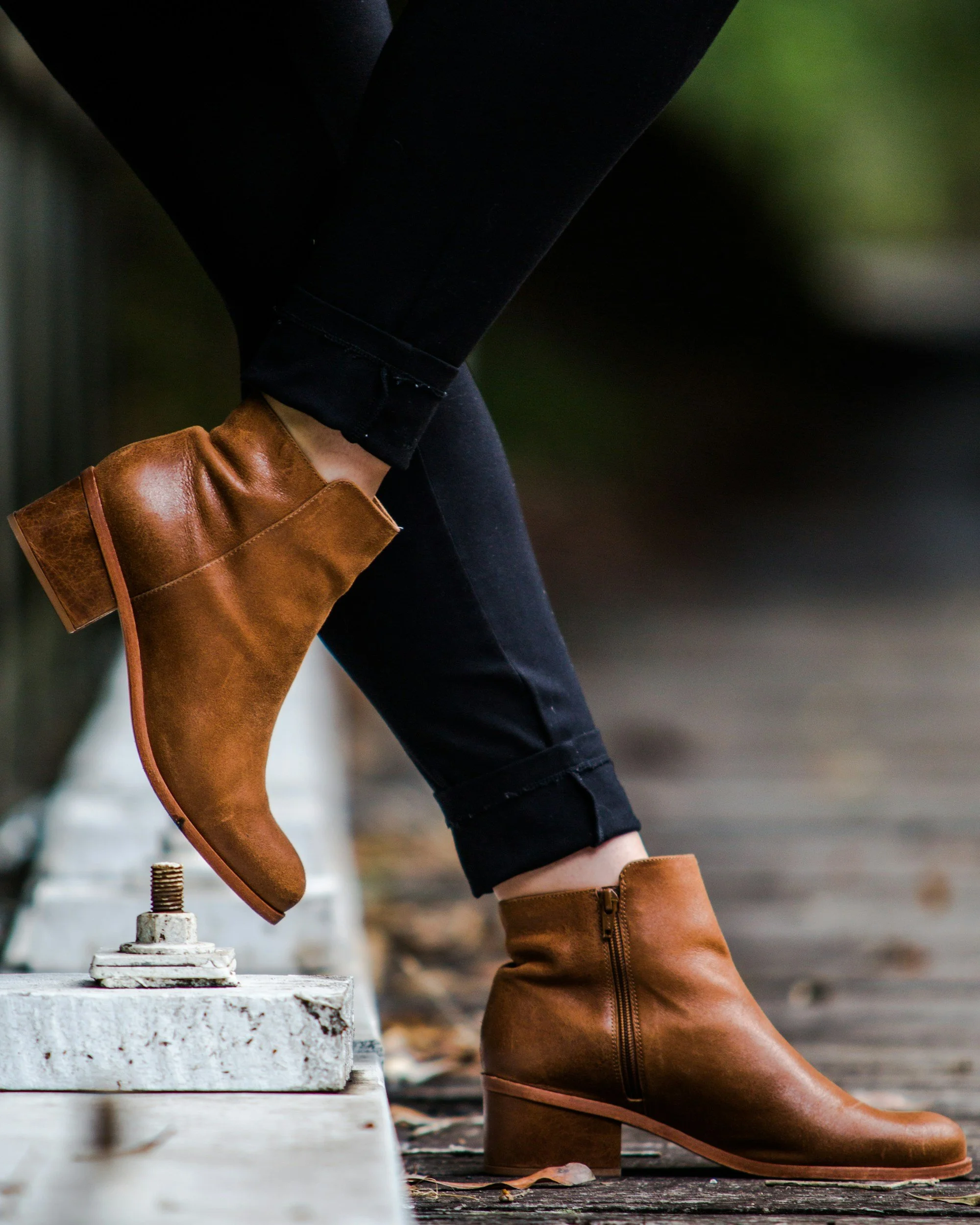 Person wearing brown leather boots and black jeans, stepping over painted white sidewalk curb, outdoor setting with blurred background.