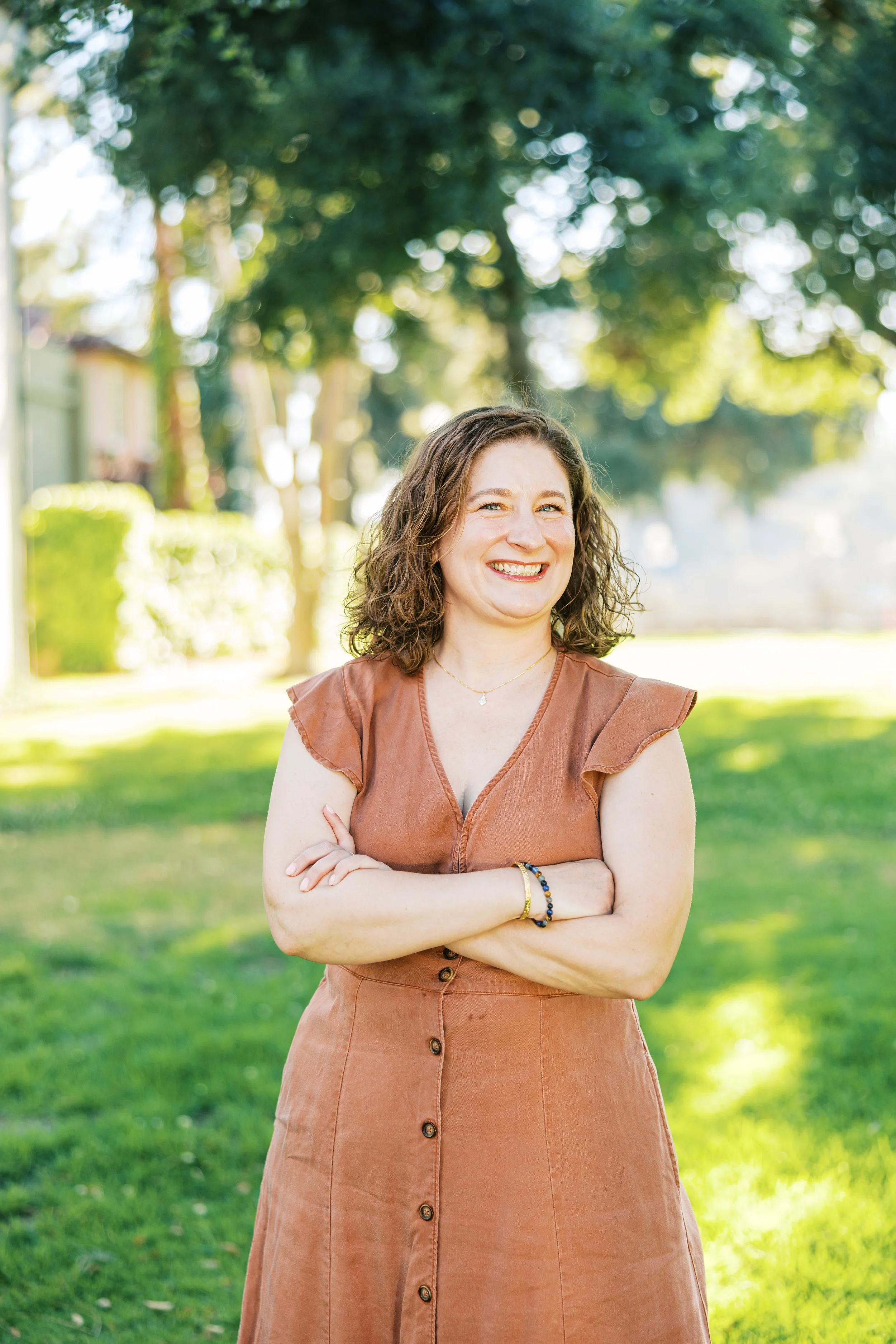 A woman with curly brown hair smiles with arms crossed, standing outdoors on a sunny day with trees and grass in the background.