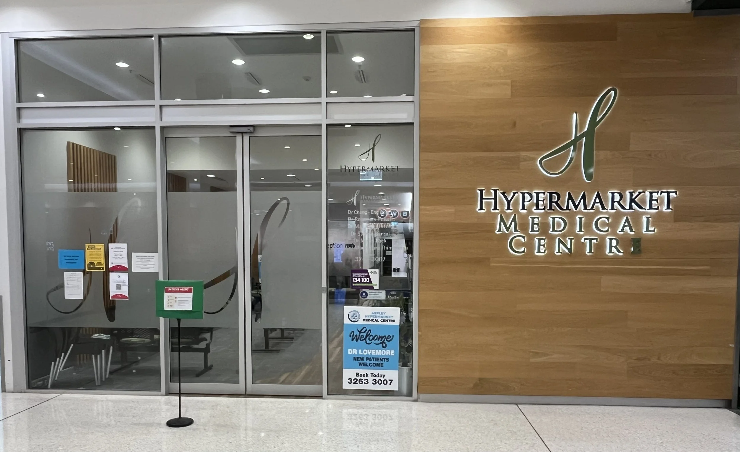 Entrance to a medical center with glass doors and a wooden wall. Sign displays 'Hypermarket Medical Center' with a logo. Notices and welcome signs on the door.