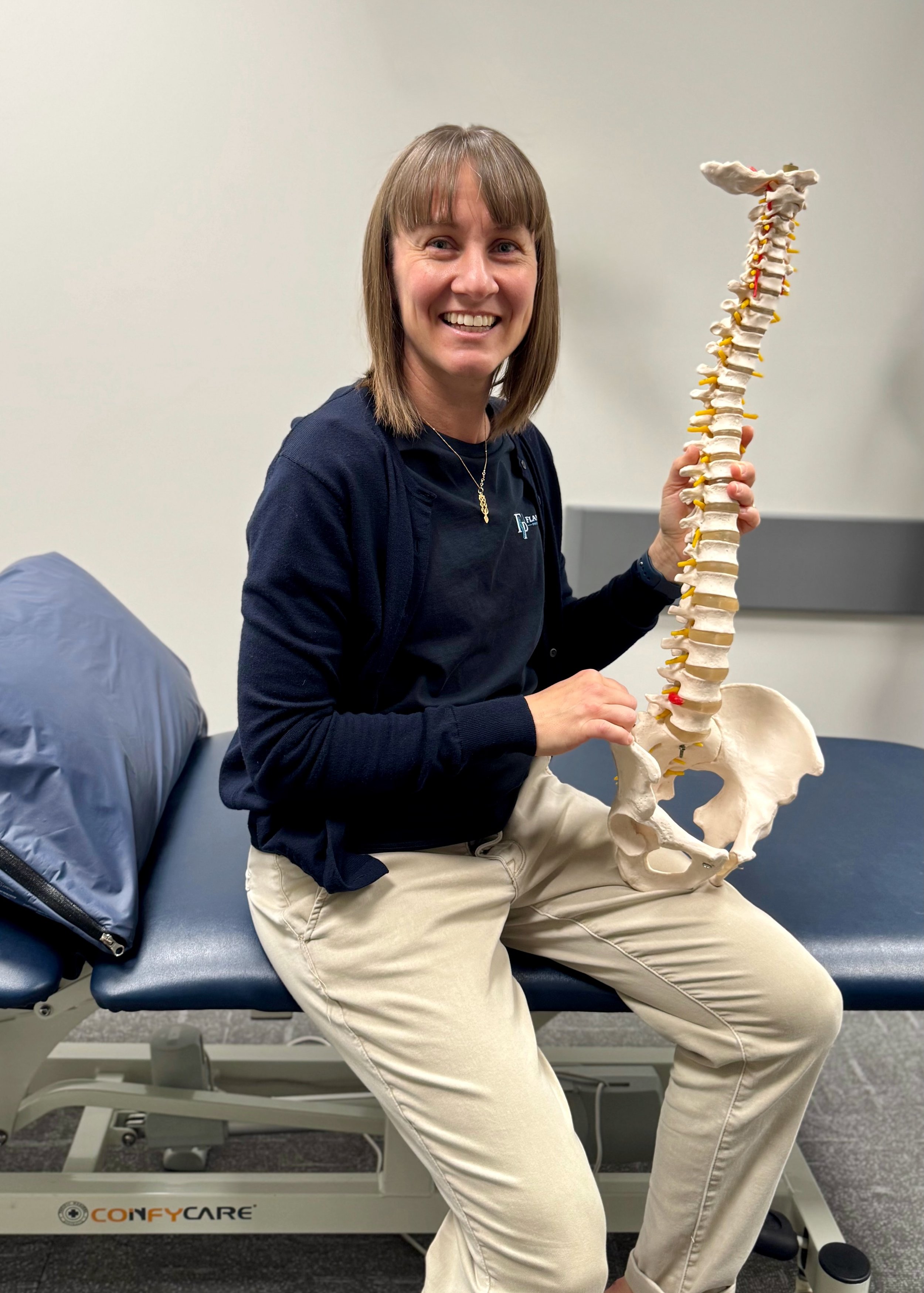 Woman sitting on a medical examination table, holding a model of a human spine, wearing a black shirt and beige pants, in a clinic or medical office.
