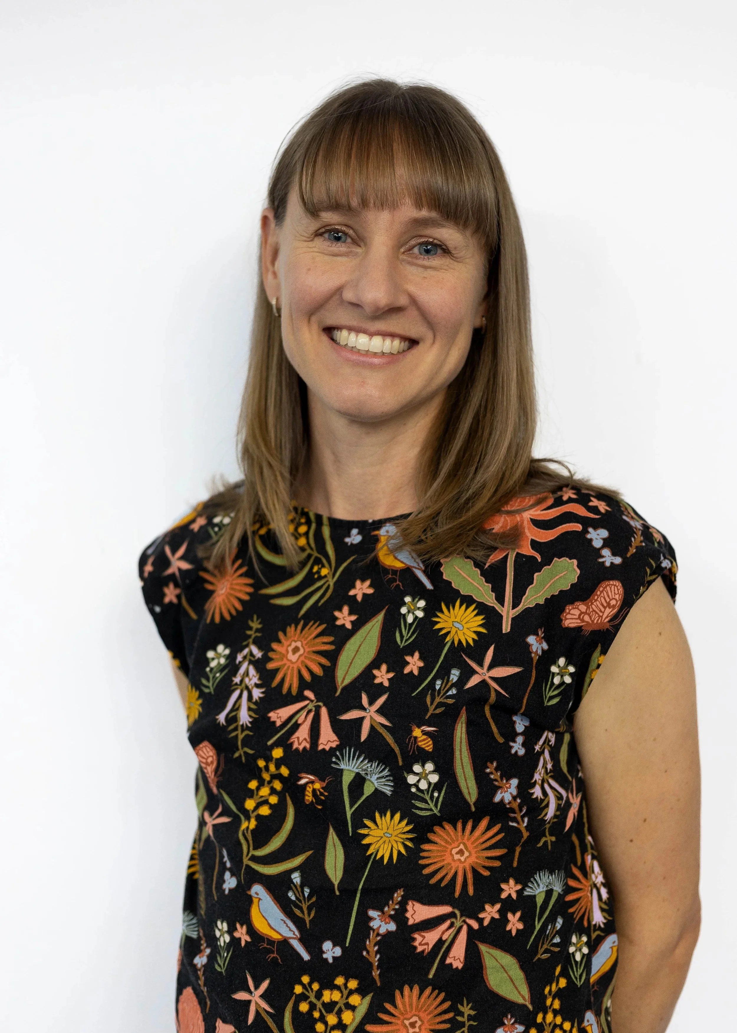 A woman physiotherapist with shoulder-length light brown hair and bangs, smiling, wearing a black floral dress with orange, yellow, white, and green flowers, standing against a plain white background.
