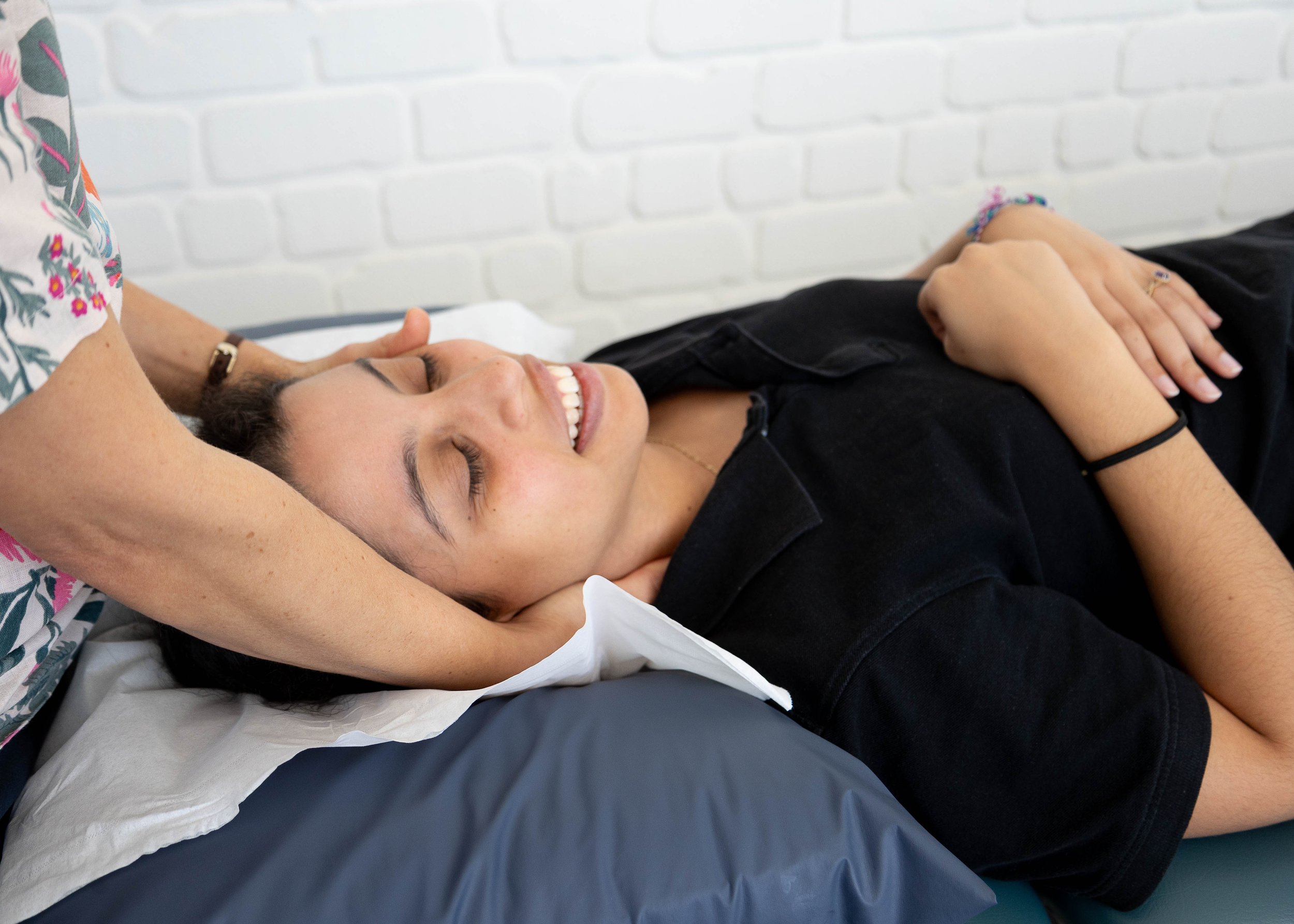 A woman with dark hair lying on her back on a bed, smiling, with her eyes closed. She has her hands resting on her chest. Physiotherapist is standing behind her treating her neck. The background shows a white brick wall.