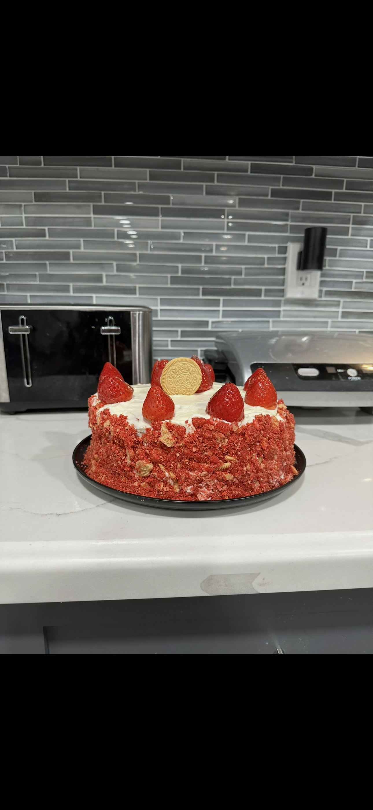 Strawberry cake on a white countertop with a gray tiled backsplash, featuring strawberries and a decorative cookie on top.