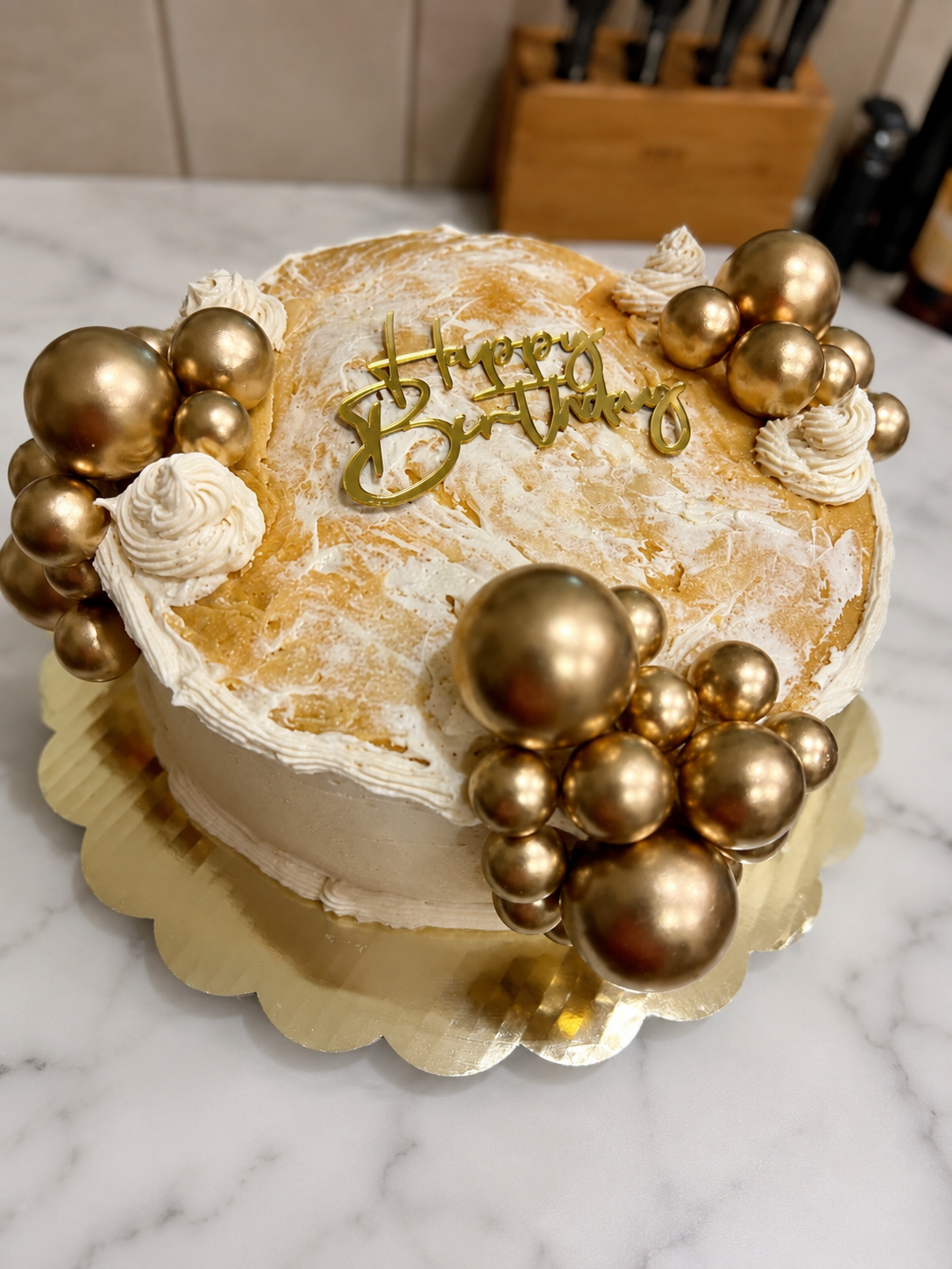 A round birthday cake decorated with gold and white elements, including gold spherical ornaments, white frosting swirls, and a gold 'Happy Birthday' topper, on a gold cake board.