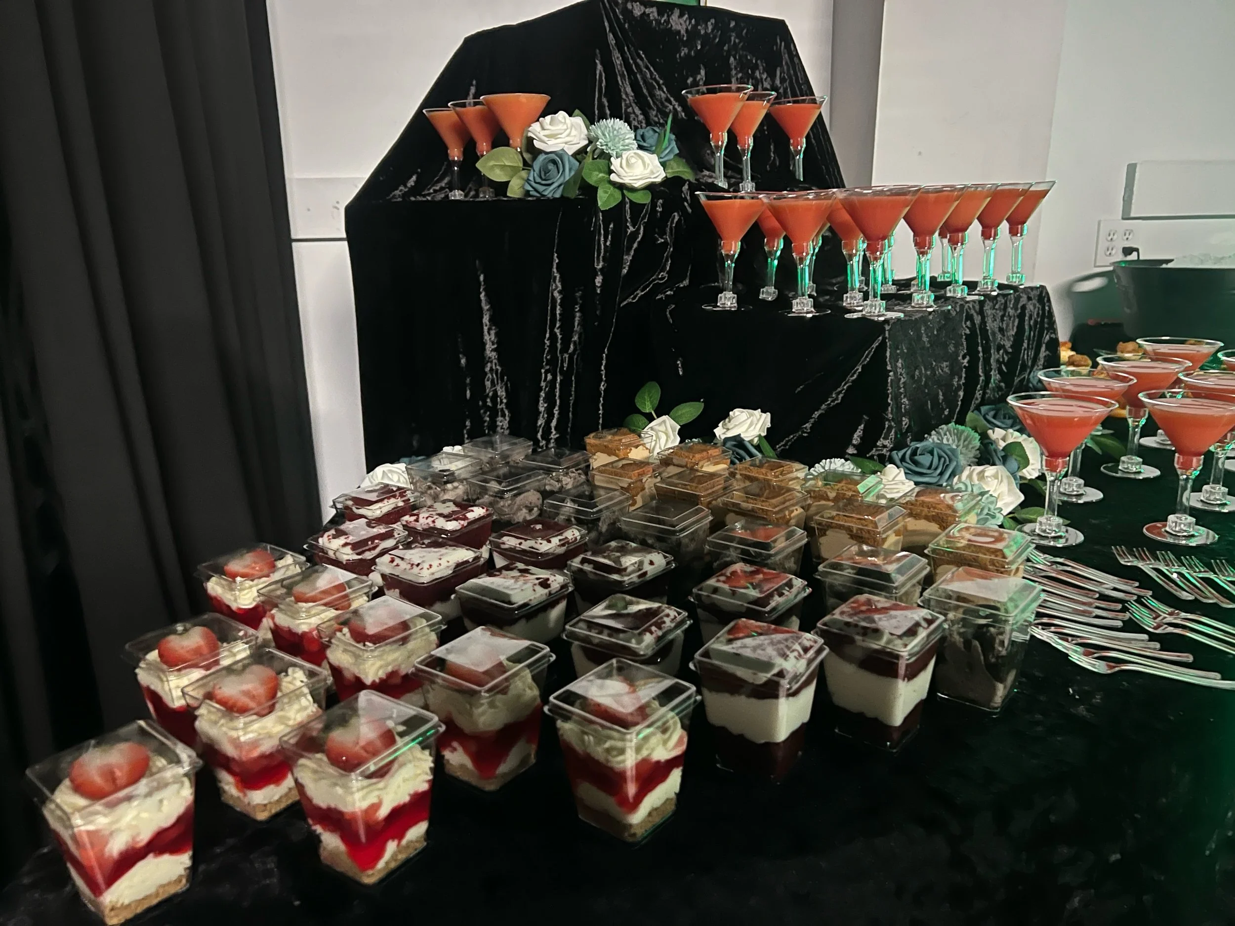 A dessert table with various cakes and layered desserts, including strawberries and cream, decorated with roses and greenery, and pink cocktails in martini glasses arranged on a black draped surface.