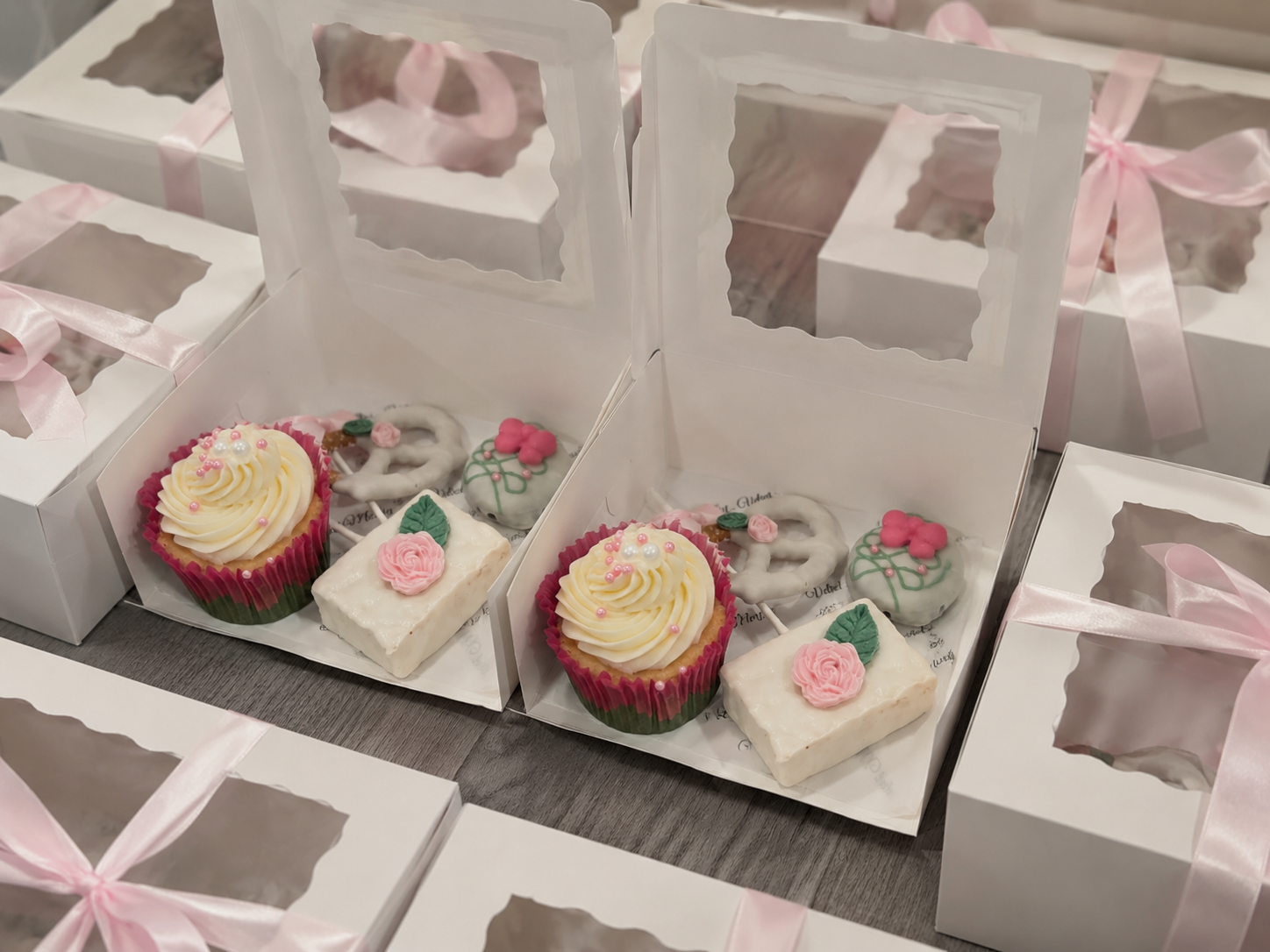 Selection of decorated cookies and cupcakes inside white gift boxes with pink ribbons, displayed on a table.