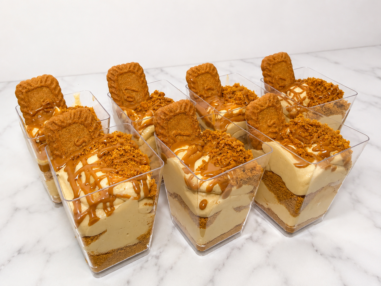 Six servings of layered dessert in clear plastic cups topped with cookies, caramel drizzle, and crushed cookies, on a white marble surface.