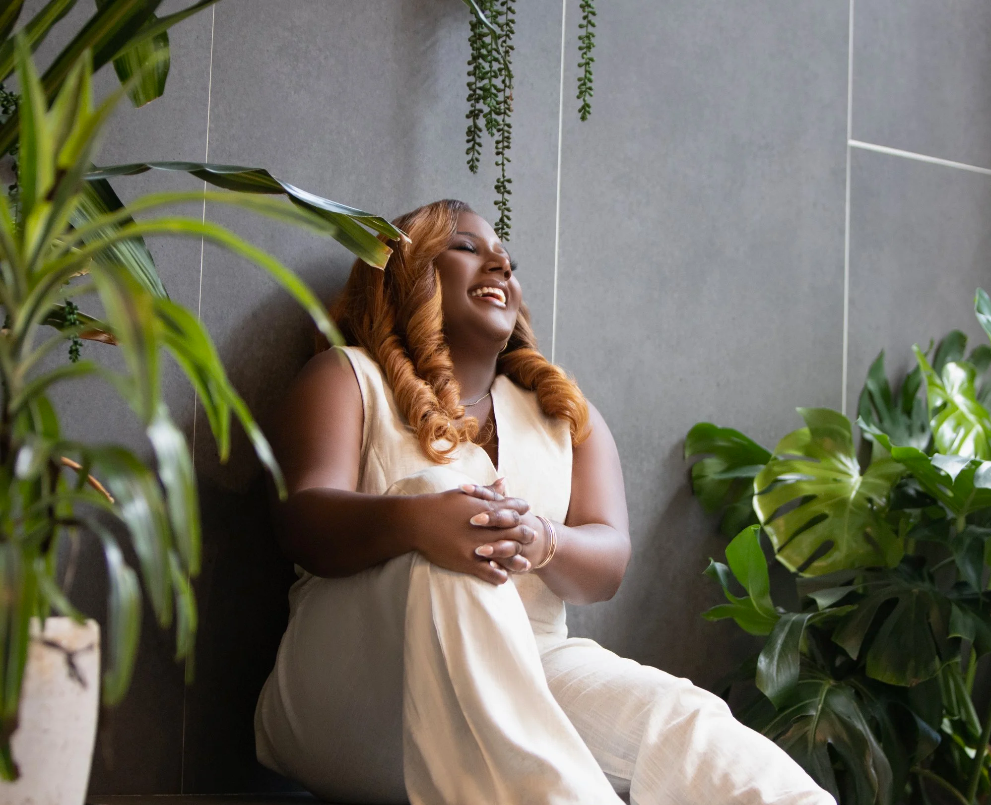 A woman with long wavy hair sitting among green plants, smiling with her eyes closed, against a gray wall.