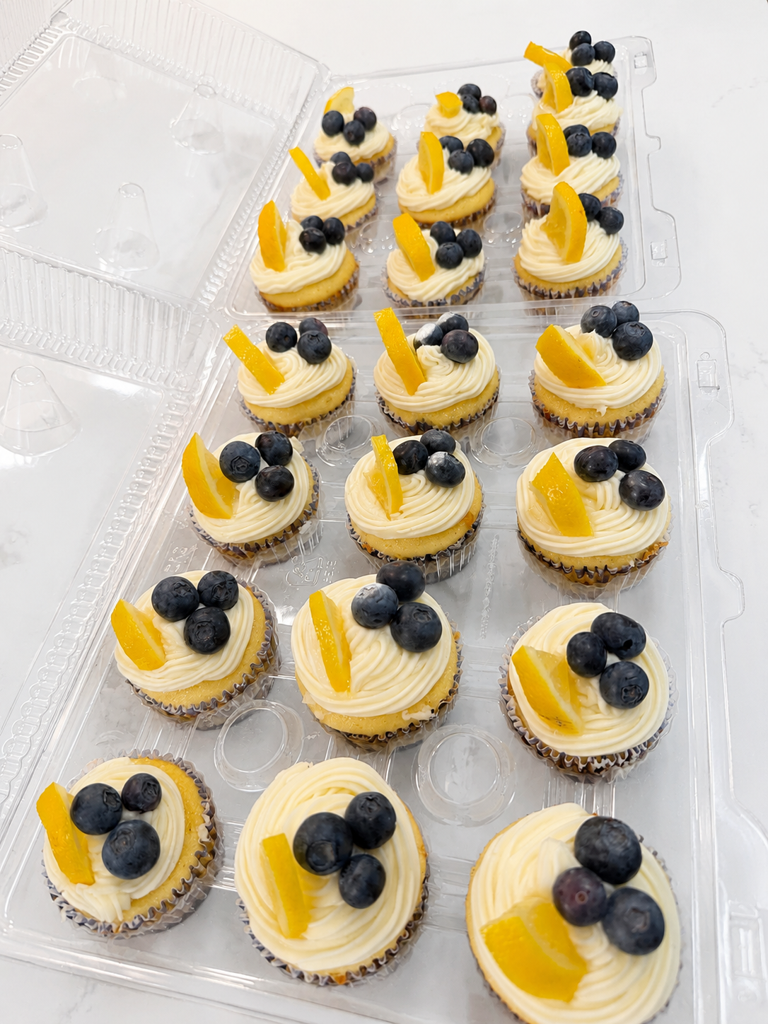 A plastic container of lemon cupcakes topped with blueberries and lemon slices.