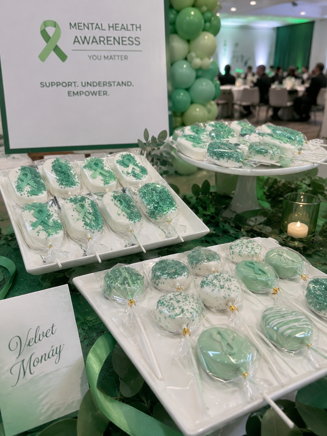Display table with green and white decorated candy apples at a mental health awareness event. The table is decorated with green balloons and a sign that reads 'Mental Health Awareness You Matter. Support. Understand. Empower.' in the background, with people sitting at tables in a conference hall.