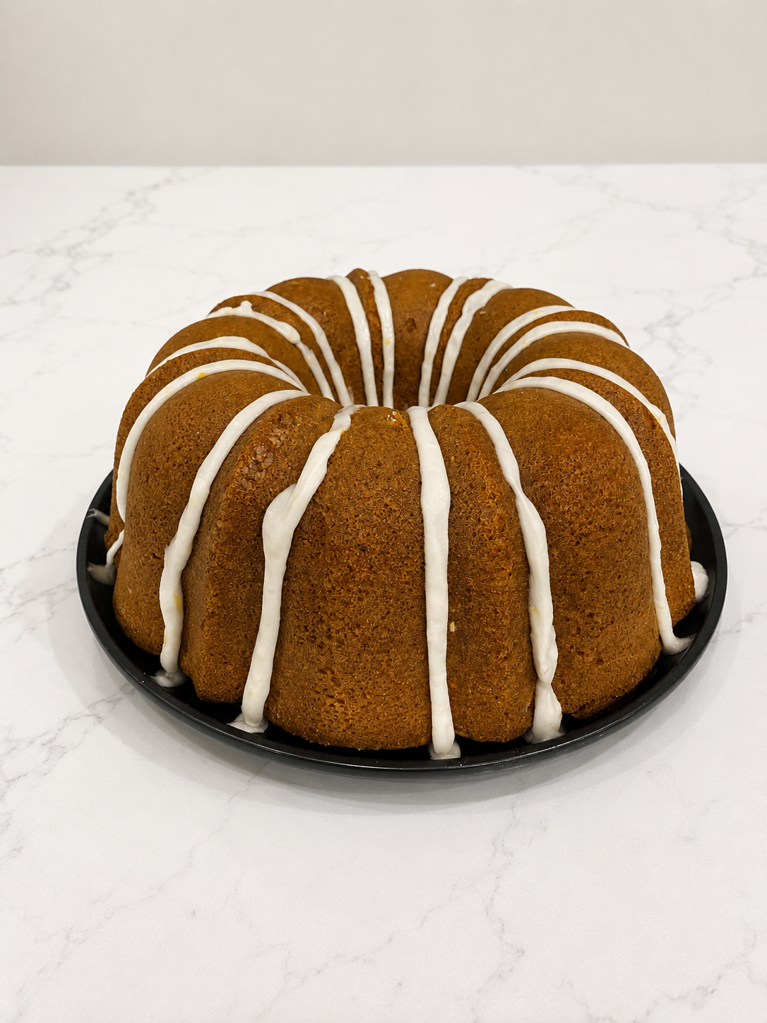 Bundt cake with white icing drizzle on black plate on white marble countertop.