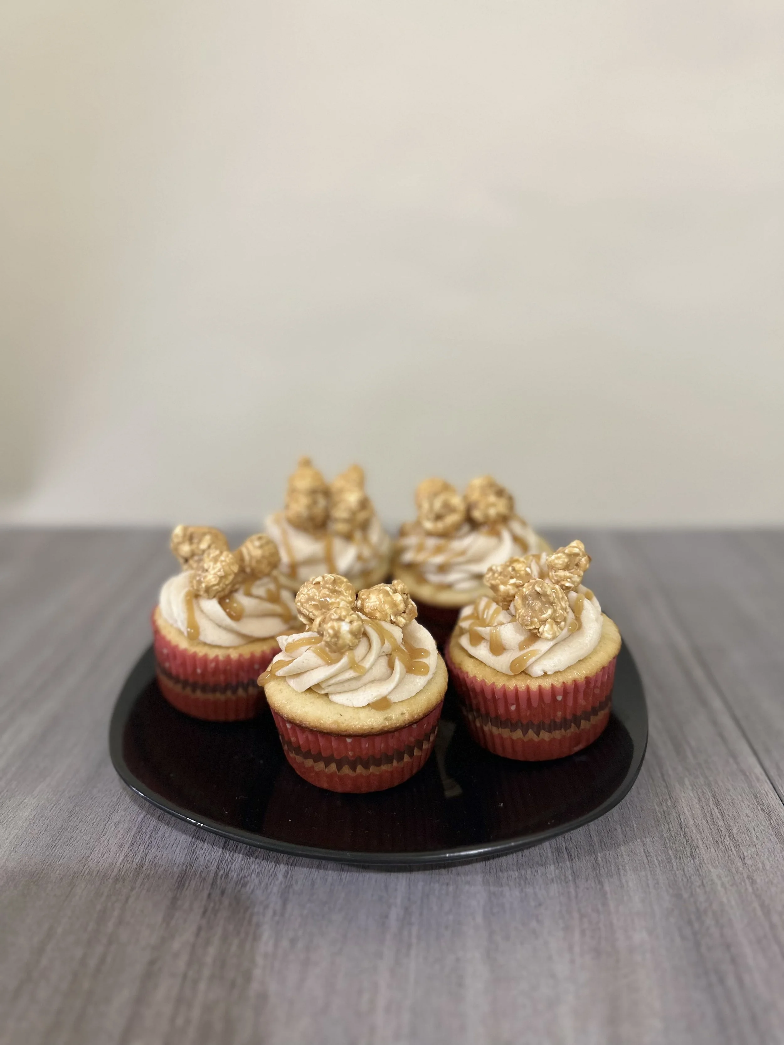 Six cupcakes with white frosting, caramel drizzle, and caramel-coated popcorn on top, placed on a black plate on a gray wooden surface.