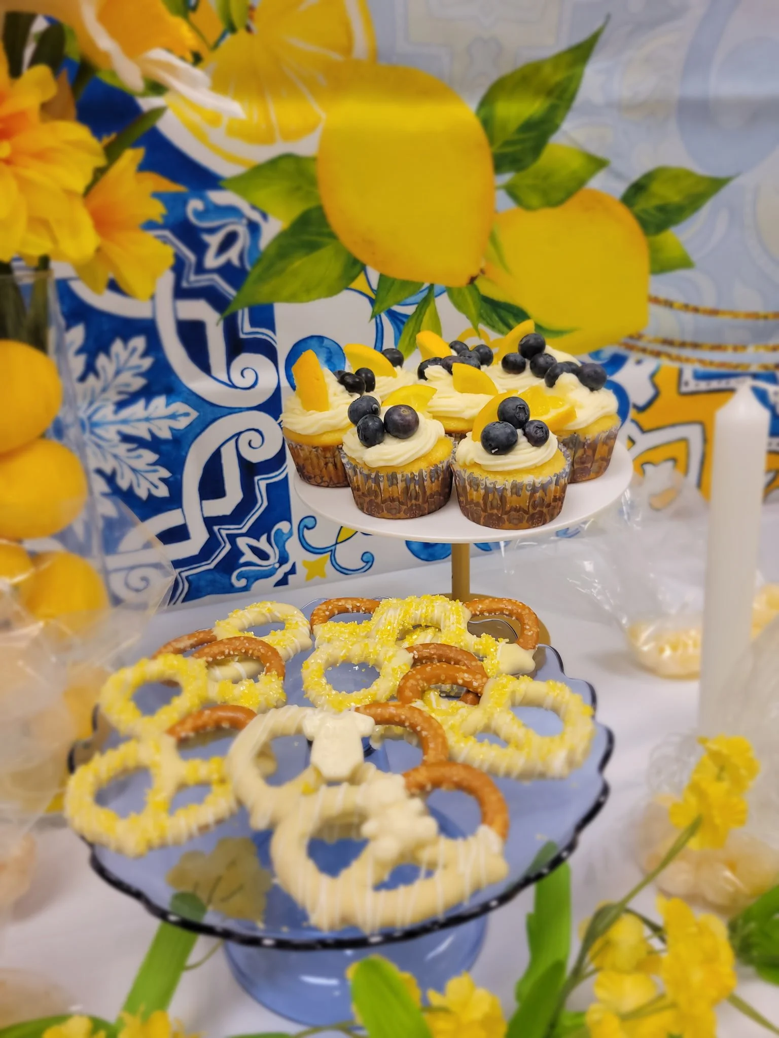 A table decorated with yellow lemons, blue and white patterned tablecloth, and a three-tiered serving stand holding lemon-themed cupcakes topped with blueberries and lemon slices, surrounded by yellow flowers.