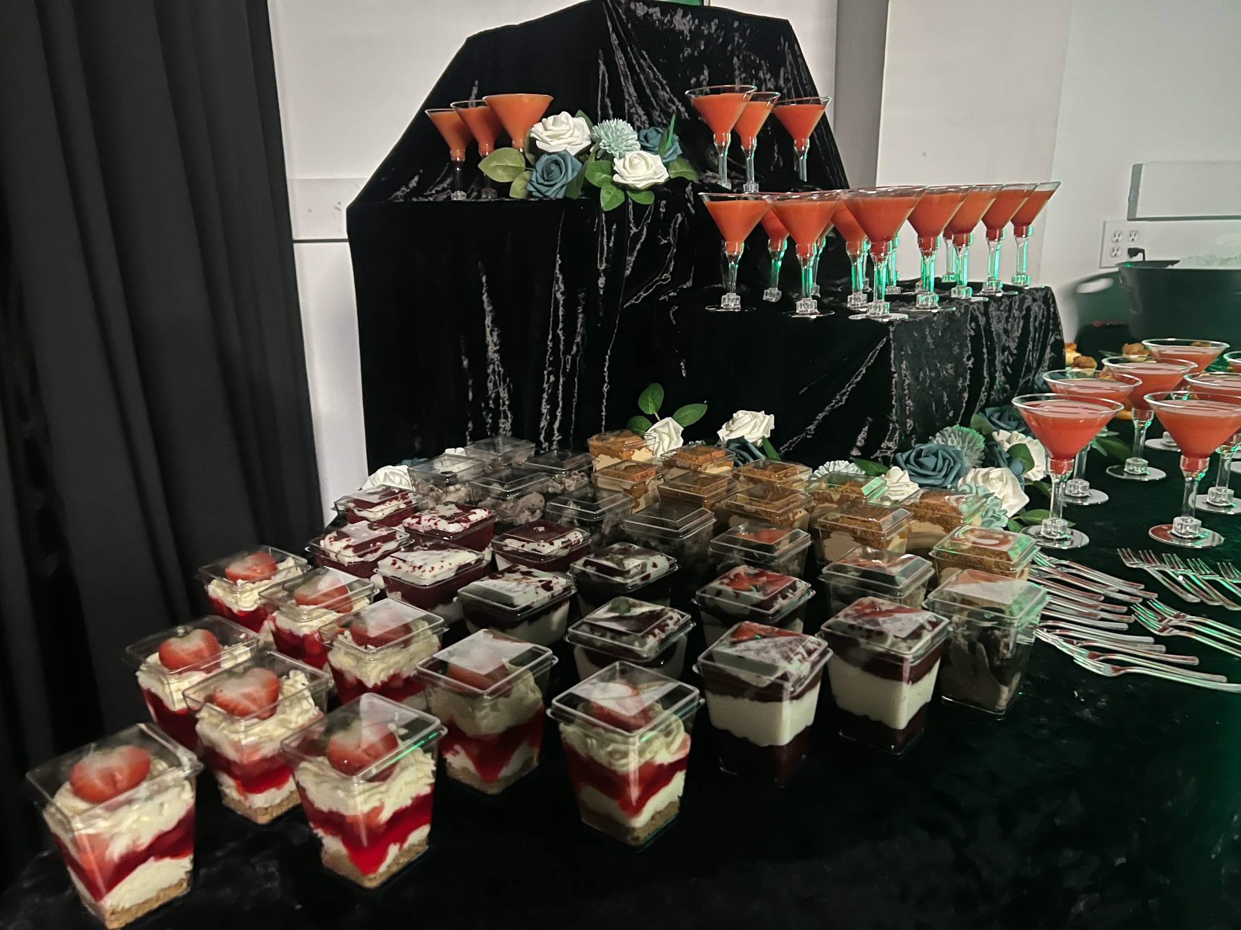 Display of numerous small dessert cups with layered strawberry and cream dessert, arranged on a black table. In the background, orange cocktails in martini glasses are arranged on a black velvet-covered stand. Decorative blue and white roses are placed among the drinks.