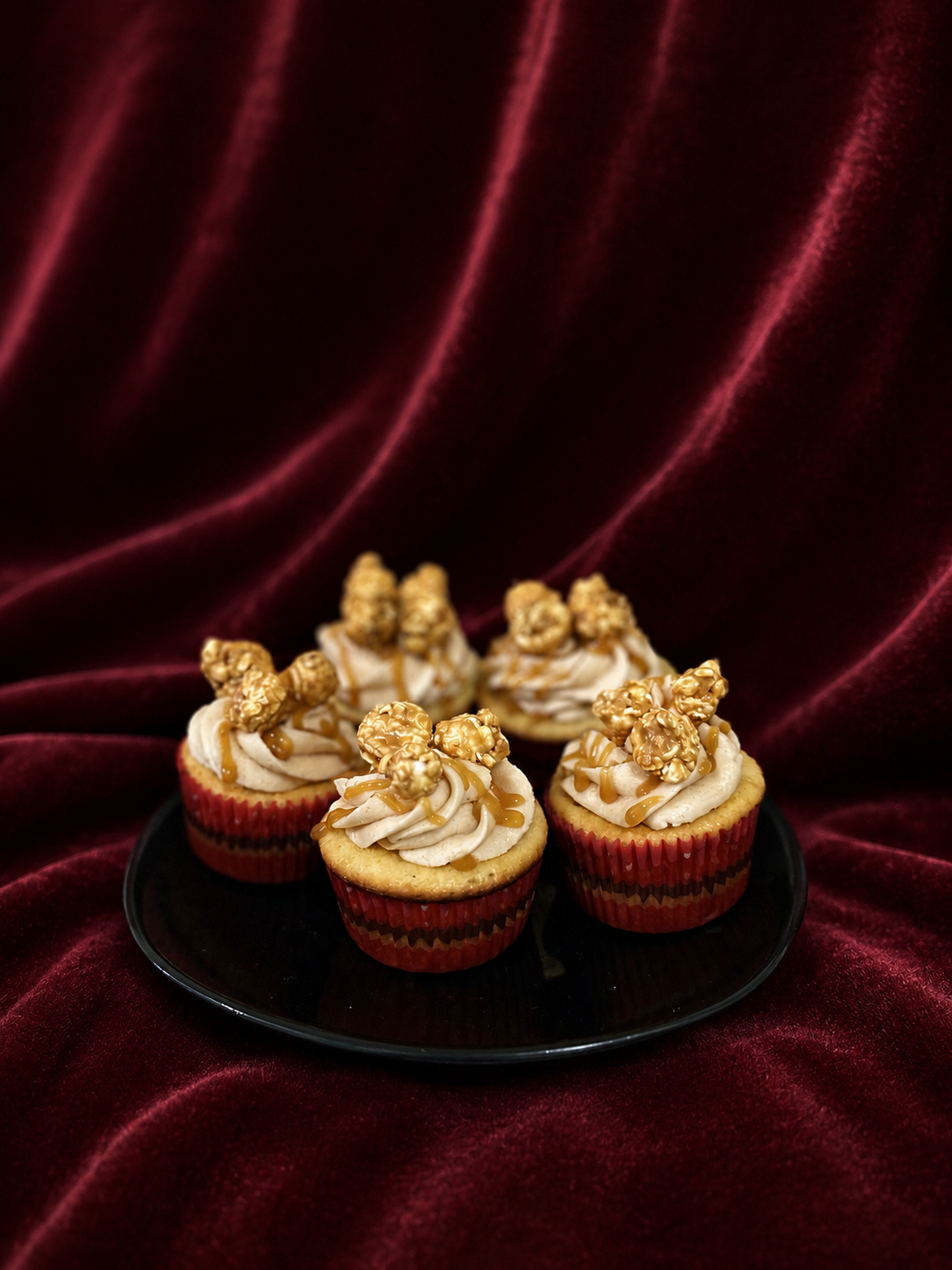 Six cupcakes with beige frosting topped with caramel drizzles and caramel-covered popcorn, arranged on a black plate against a dark red velvet background.