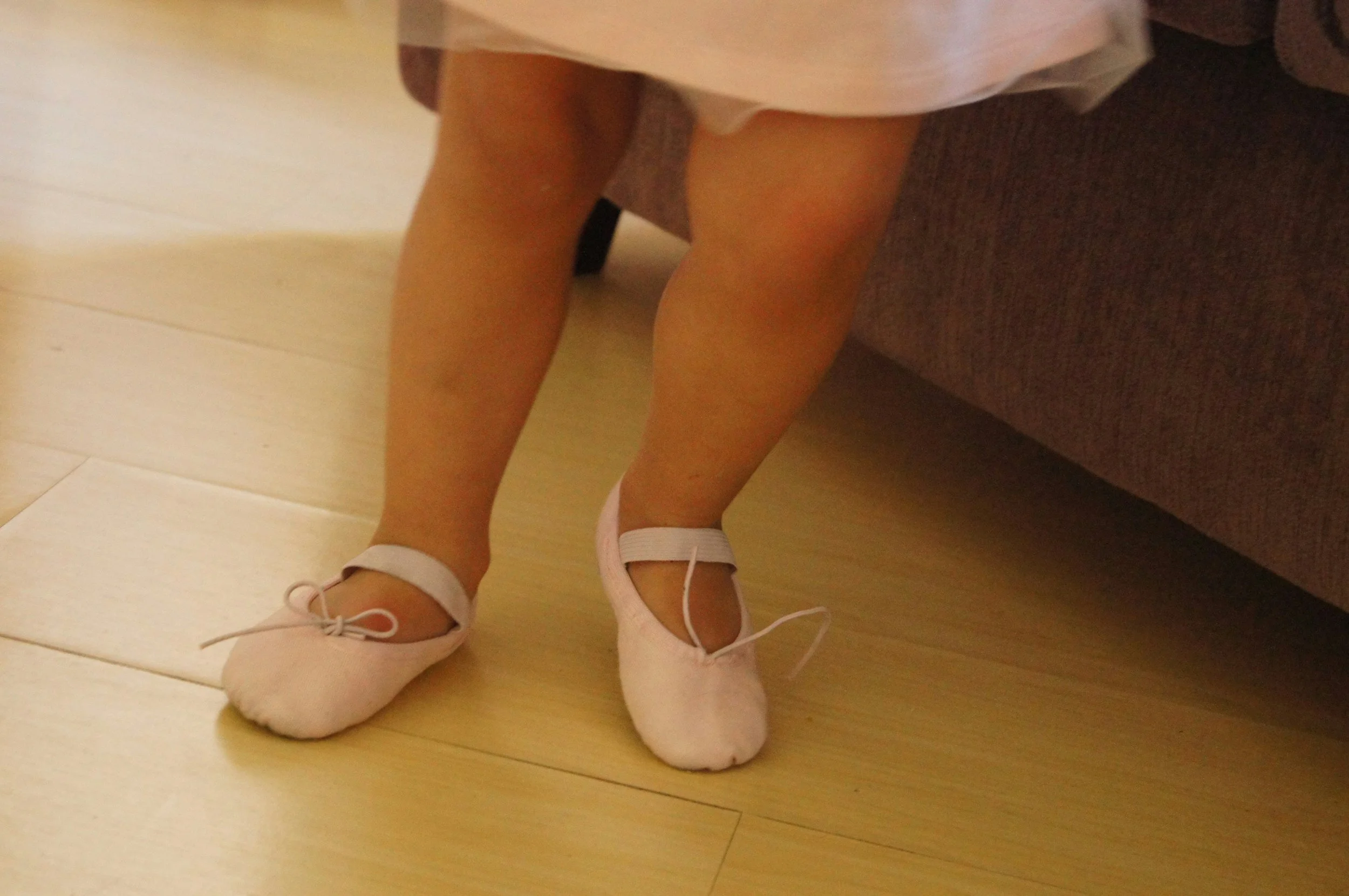 Close-up of a child's legs wearing pink ballet shoes, standing on a wooden floor near a brown couch.