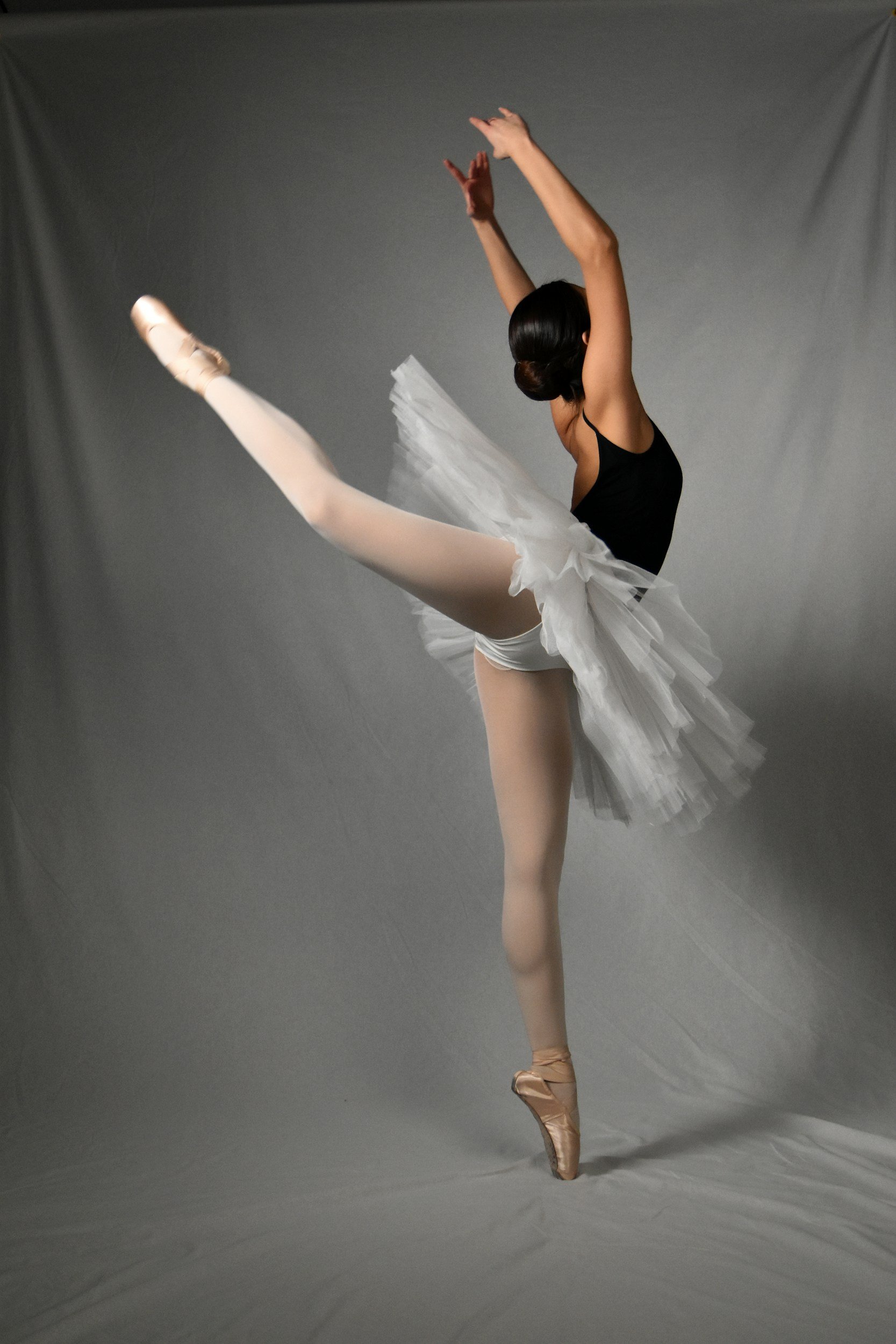 A ballet dancer performing a graceful leap, wearing a white tutu, pink pointe shoes, and a black leotard, with her arms raised above her head against a plain gray background.