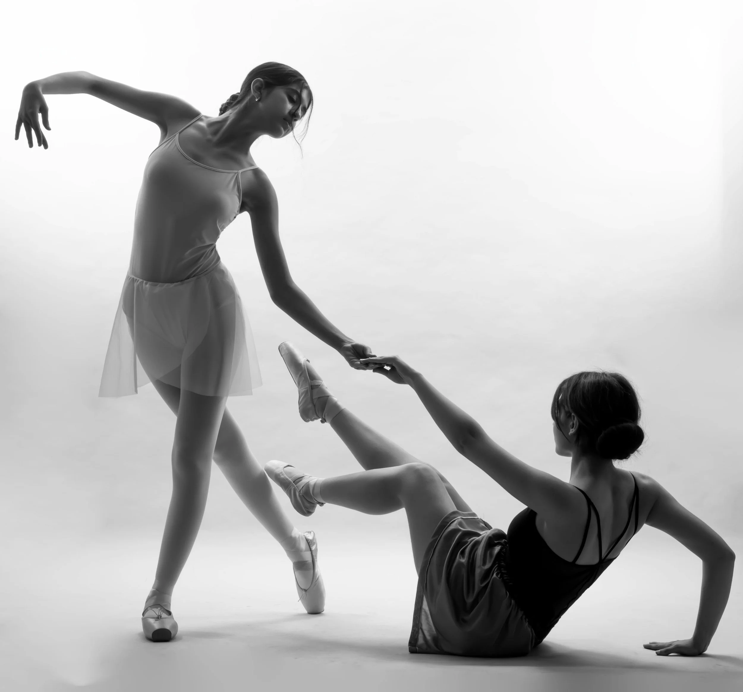 Black-and-white photo of a ballerina performing a dance pose, supported by another dancer lying on the ground, with a plain background.