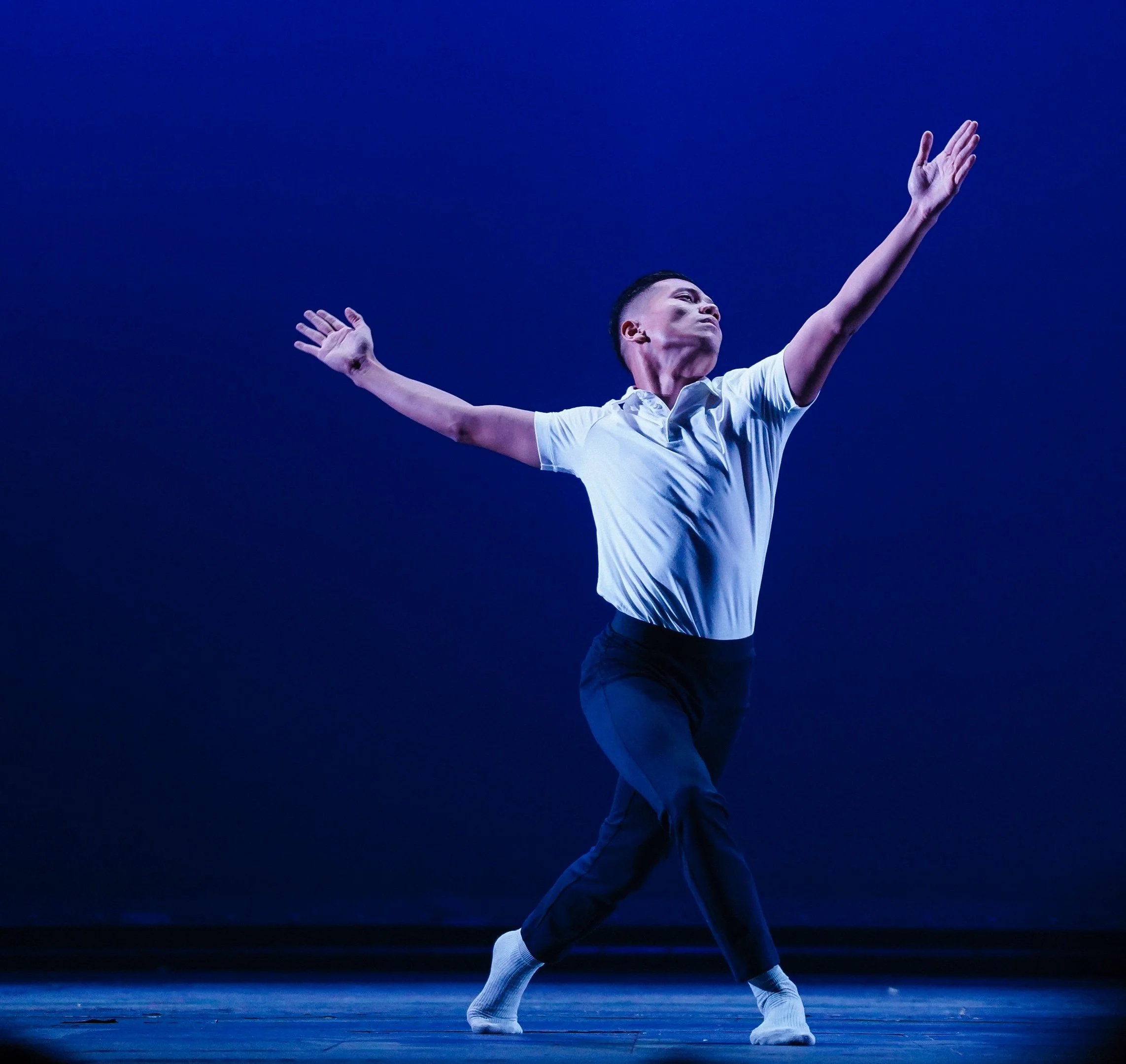 Naim performing on stage with arms extended and one leg lifted, wearing a white shirt and dark pants, against a dark blue background.