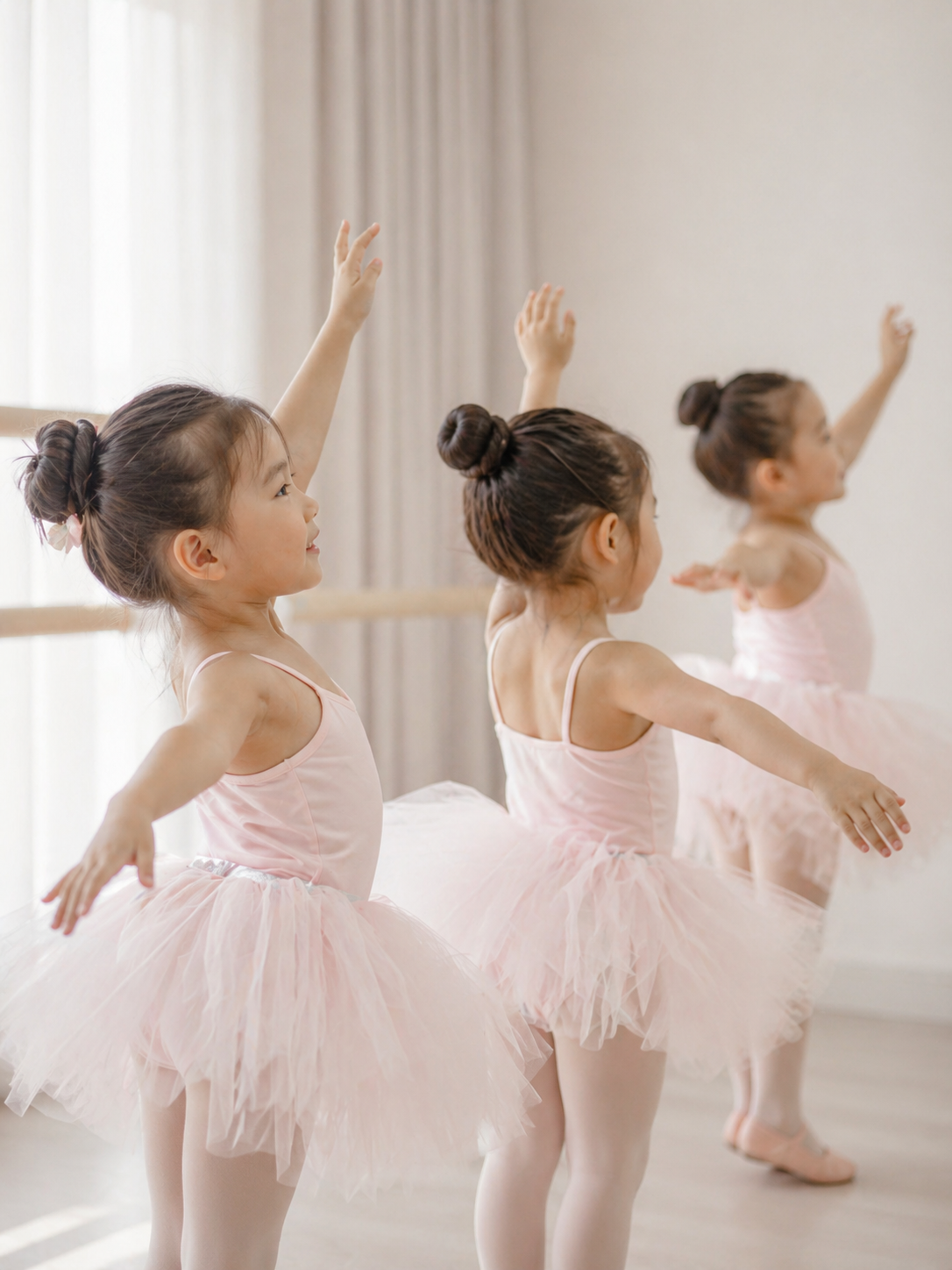 Three young girls in pink ballet tutus practicing ballet in a dance studio, raising arms in a ballet pose.