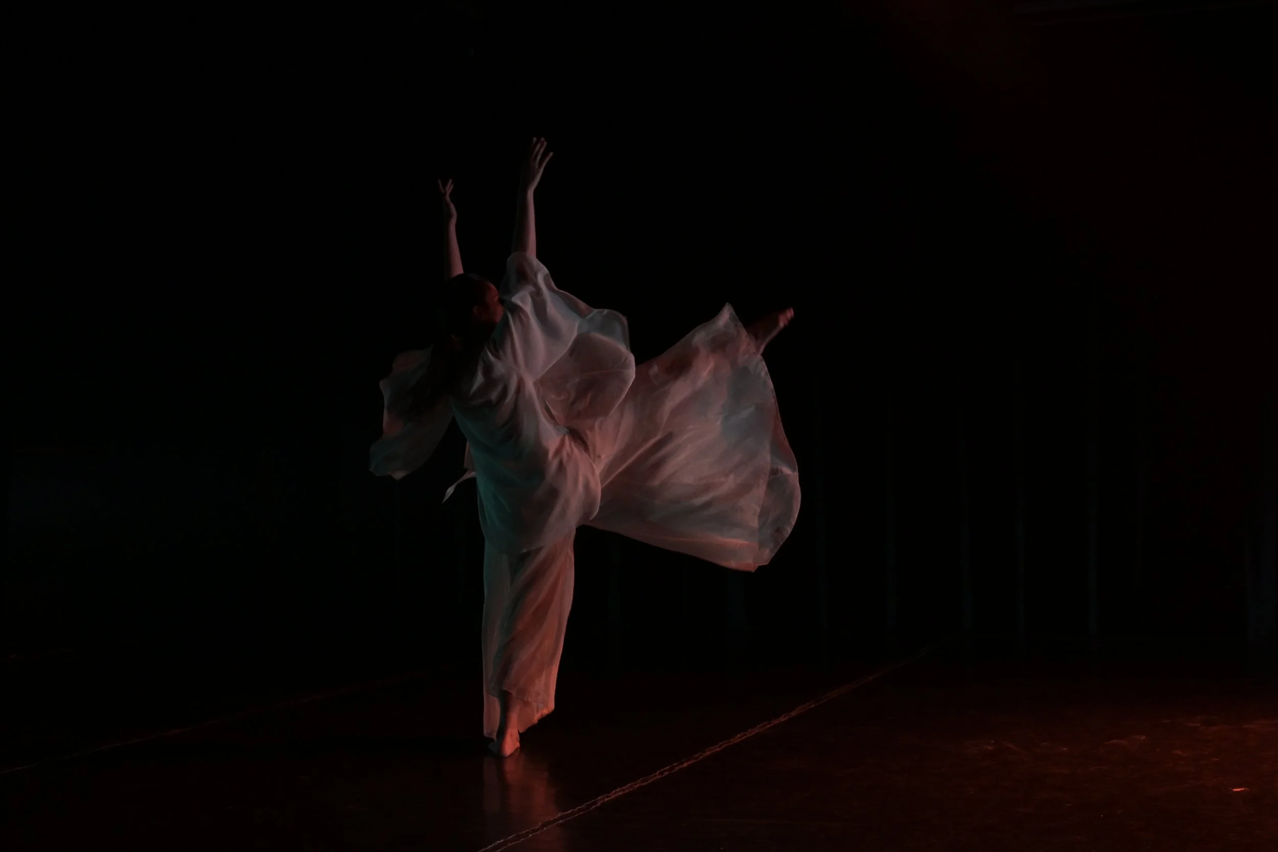 A person in a flowing dress is performing a dance or pose on a dark stage, with arms extended and backdrop absent.