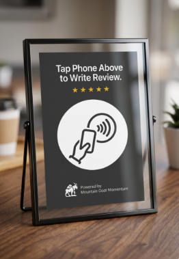 Framed sign on a wooden table instructing customers to tap phone above to write review, with a hand holding a phone icon and sound wave symbol, and five stars underneath.