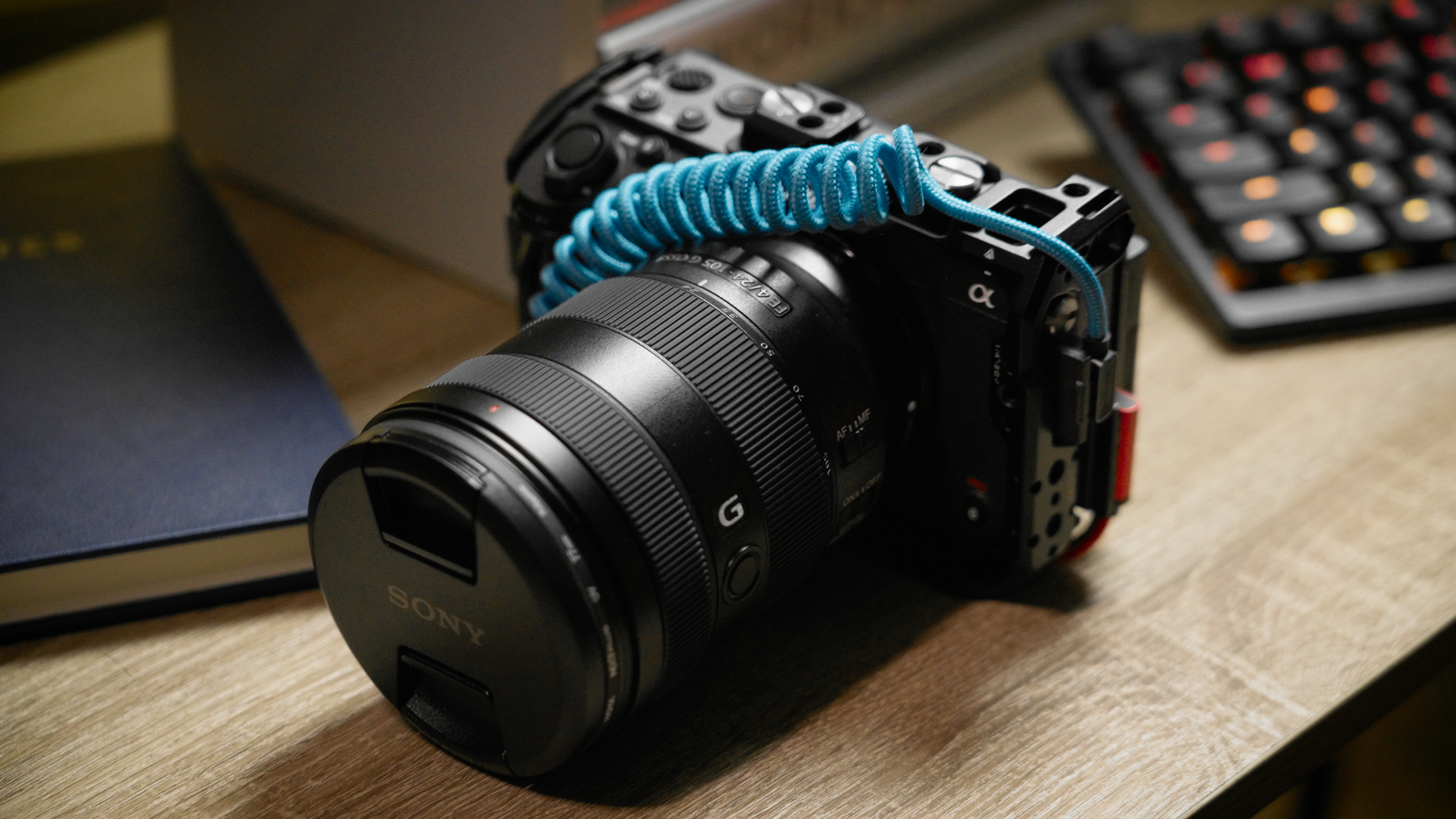 A Sony camera with a blue coiled strap placed on a wooden surface, with a calculator and a black object nearby.
